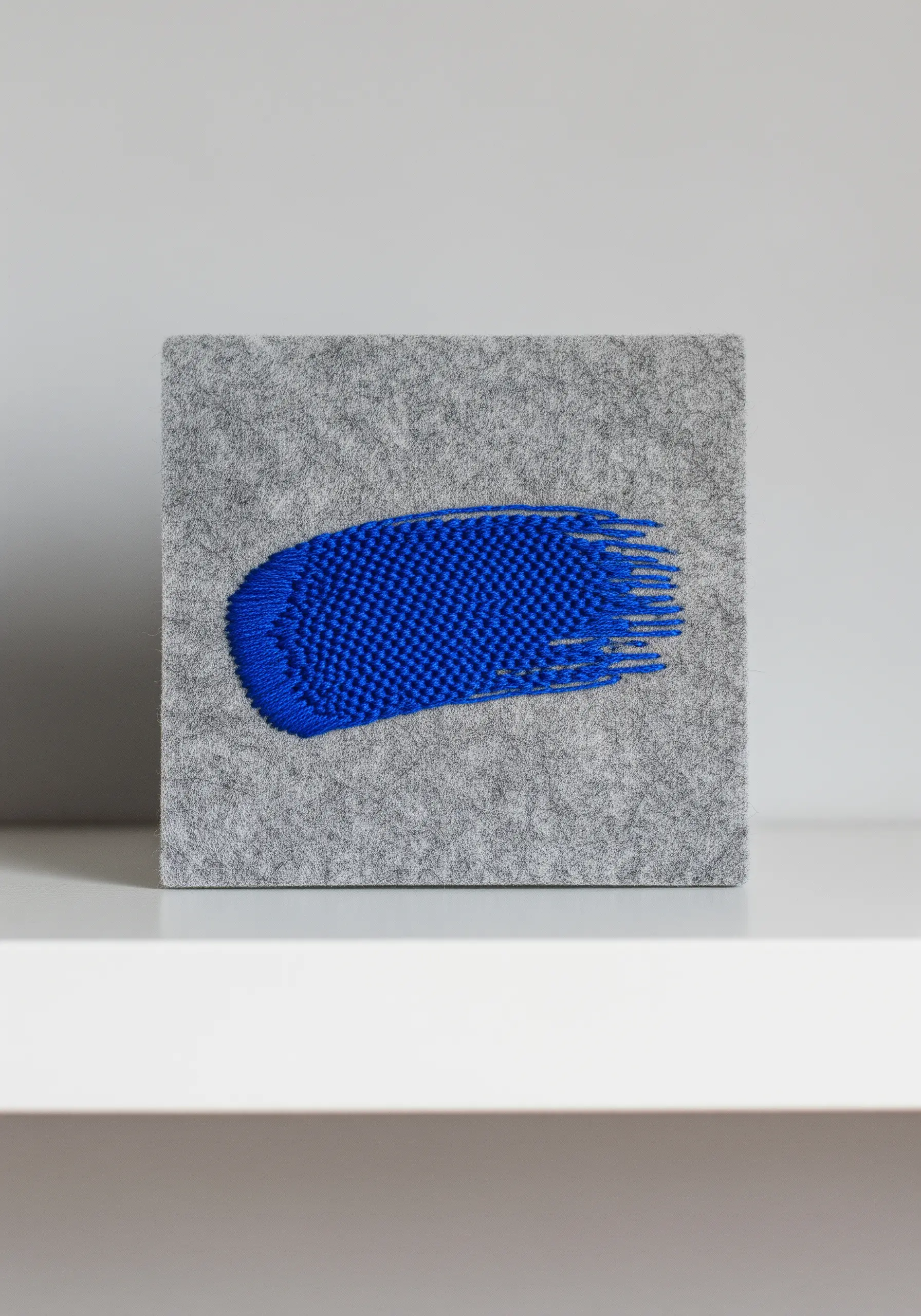 A bold blue, textured paint-stroke motif embroidered on a square of gray felt.