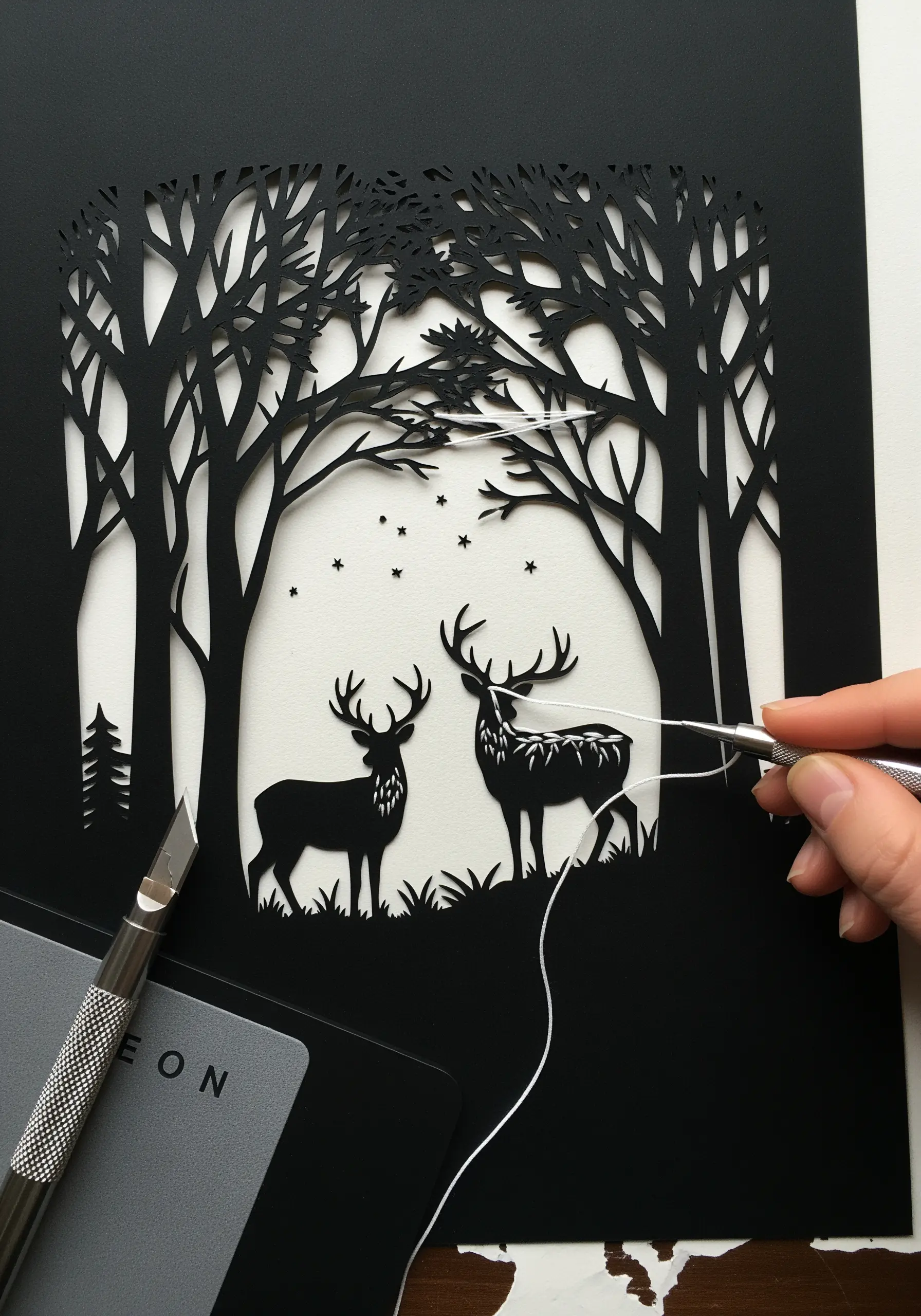 A delicate papercut forest scene with white thread being stitched onto a deer silhouette.