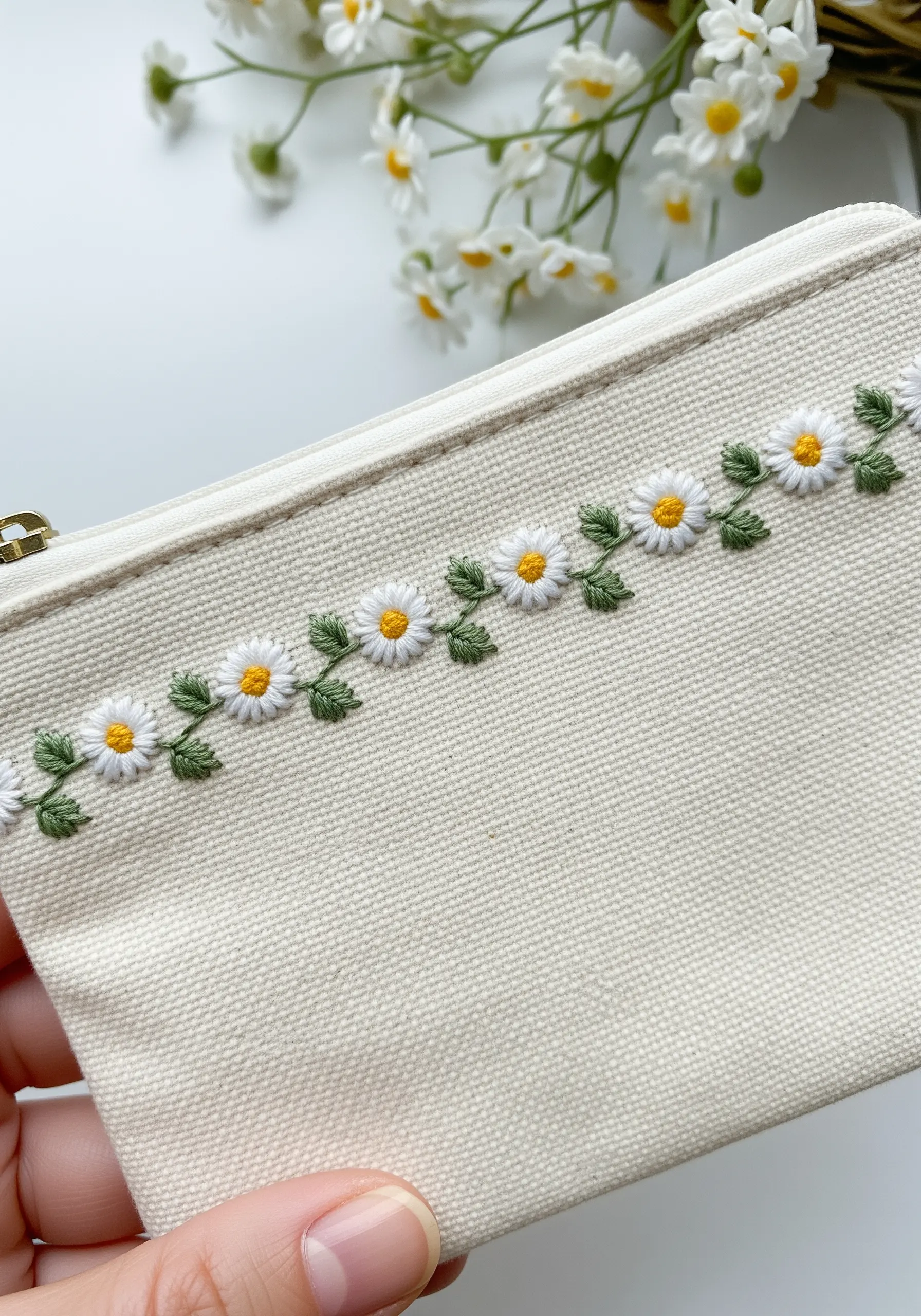 A neat border of tiny, uniform daisies and leaves on a canvas zipper pouch.