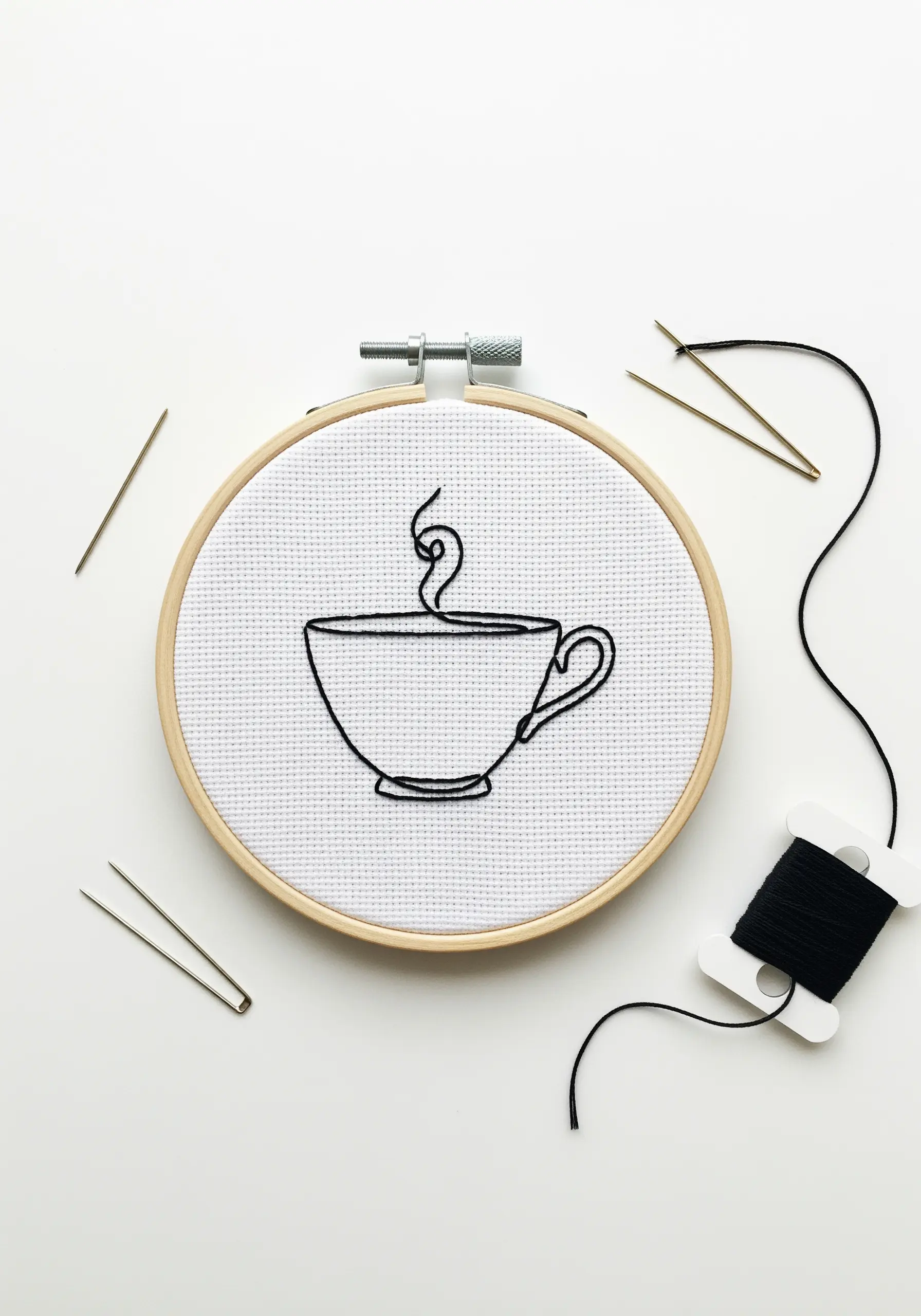 Clean line art of a coffee cup embroidered in black thread on white Aida cloth.