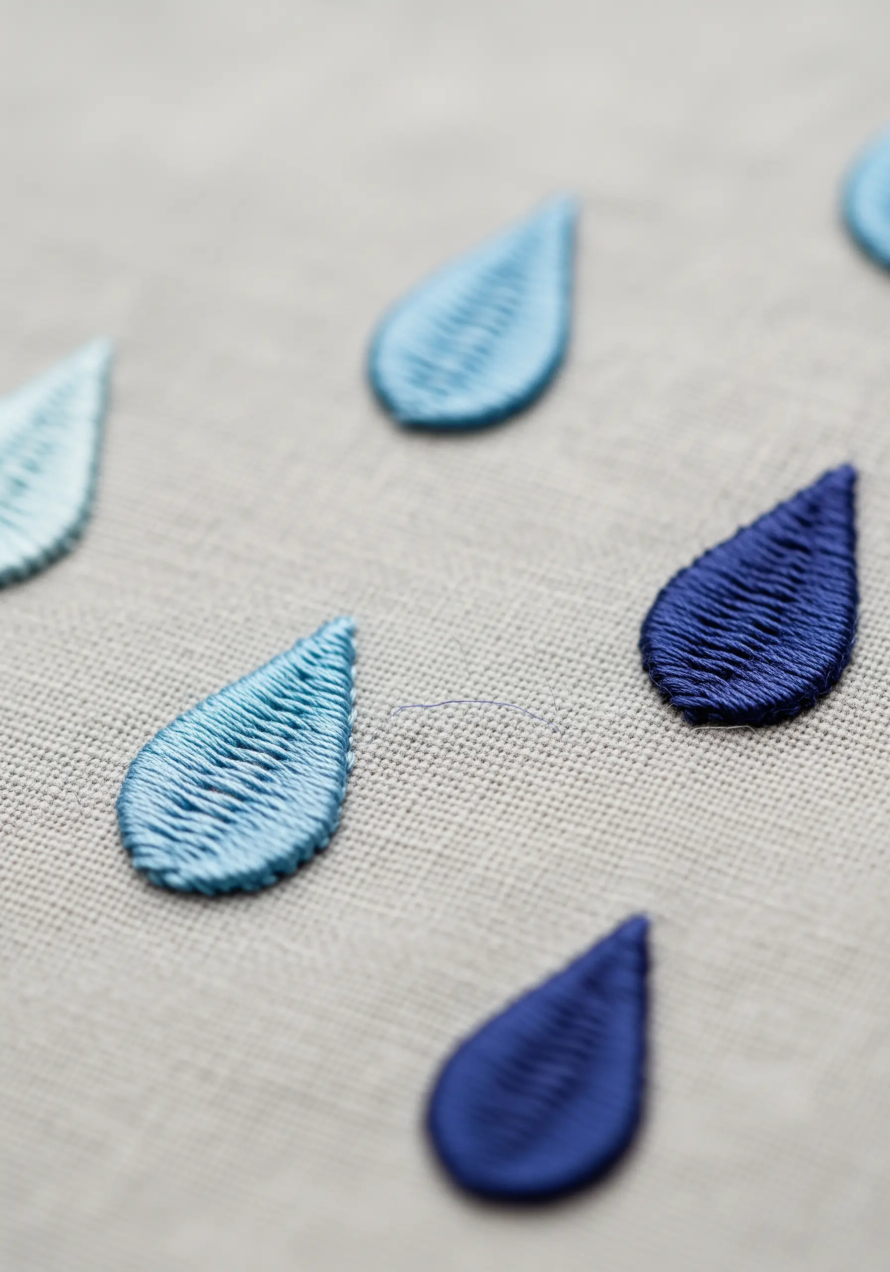 Several satin-stitched raindrops in varying shades of blue on a gray linen background.