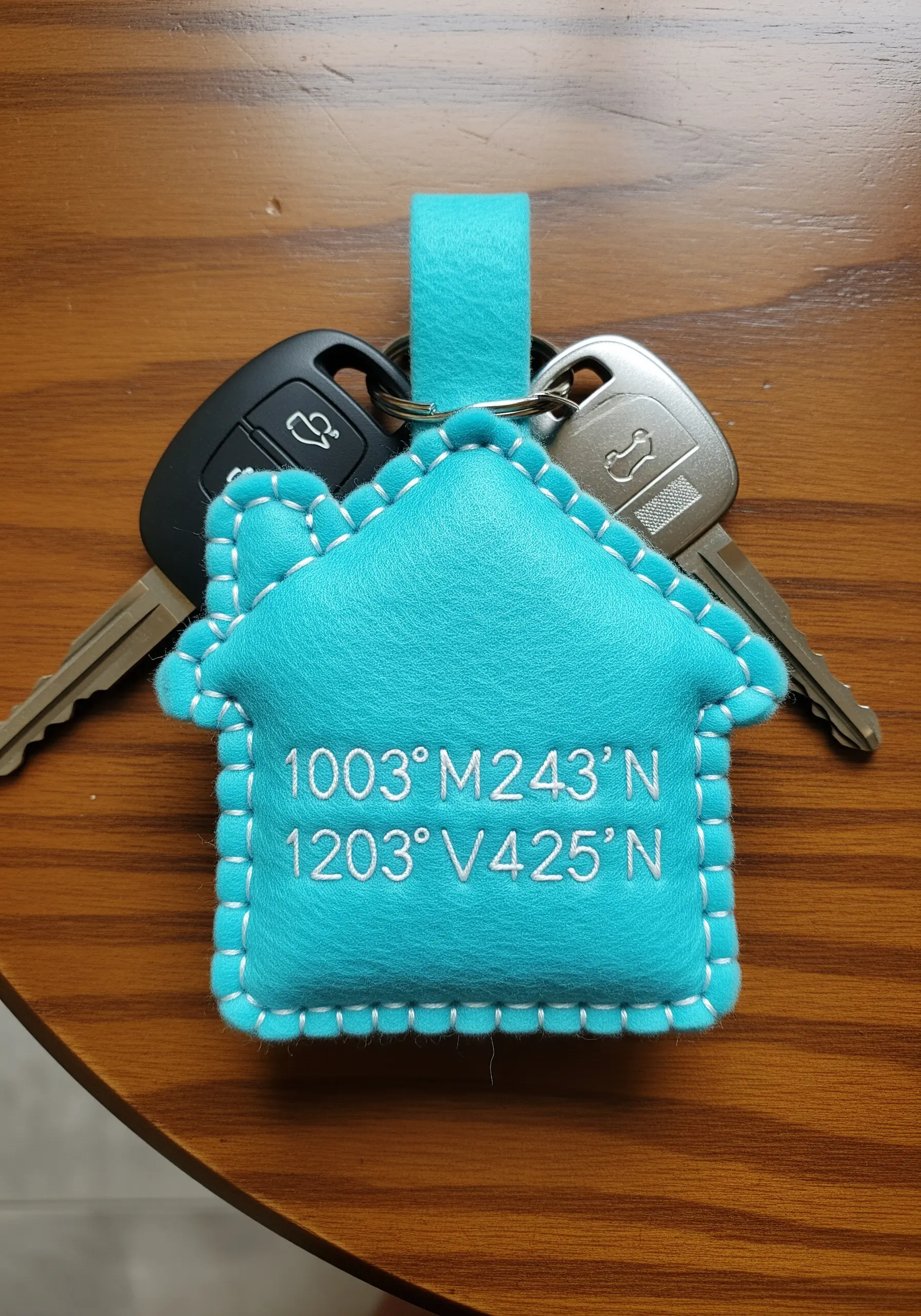 A house-shaped felt keychain in turquoise, embroidered with white GPS coordinates.