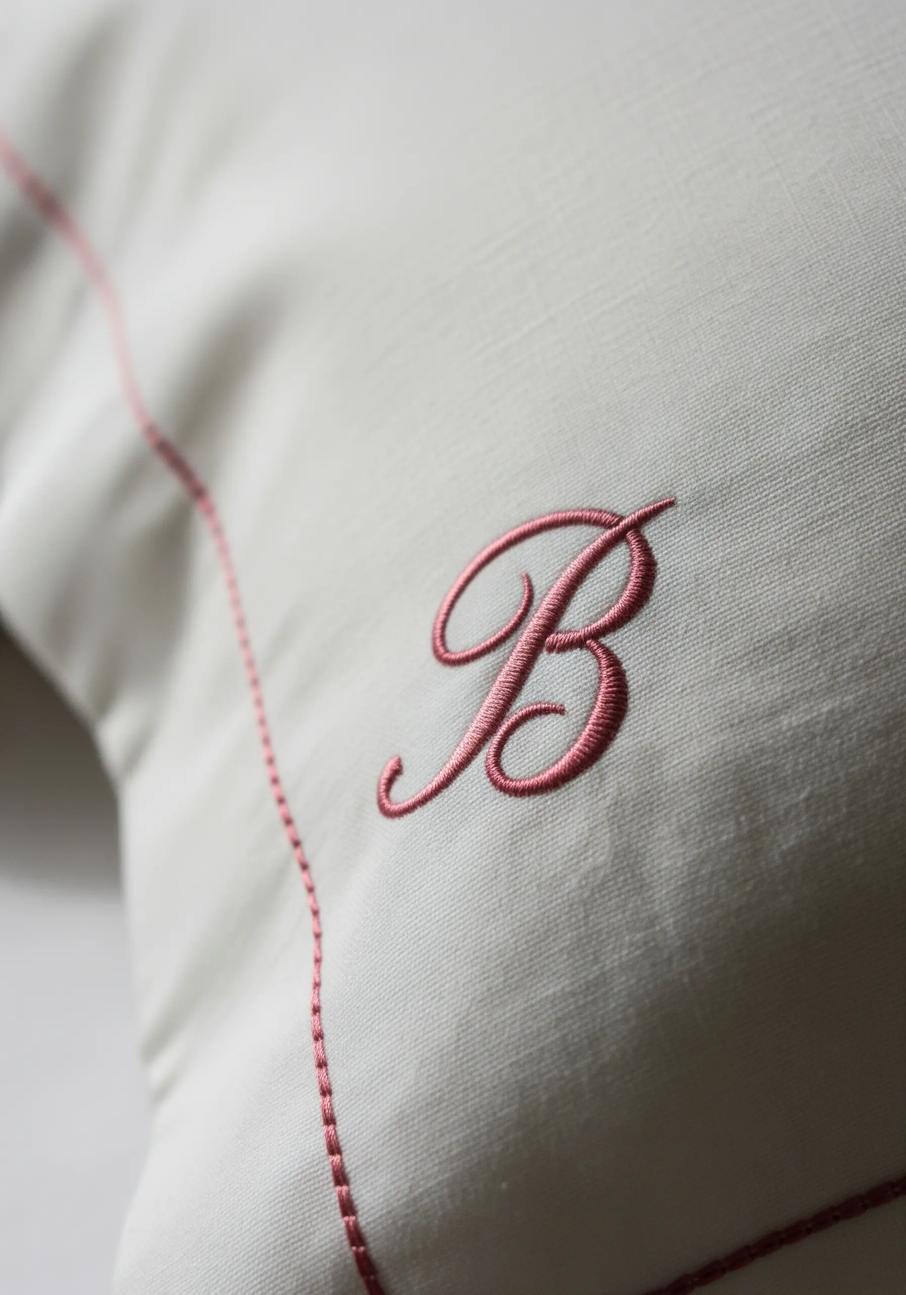 A pink, cursive letter 'B' monogram embroidered on an off-white pillowcase with a matching border.