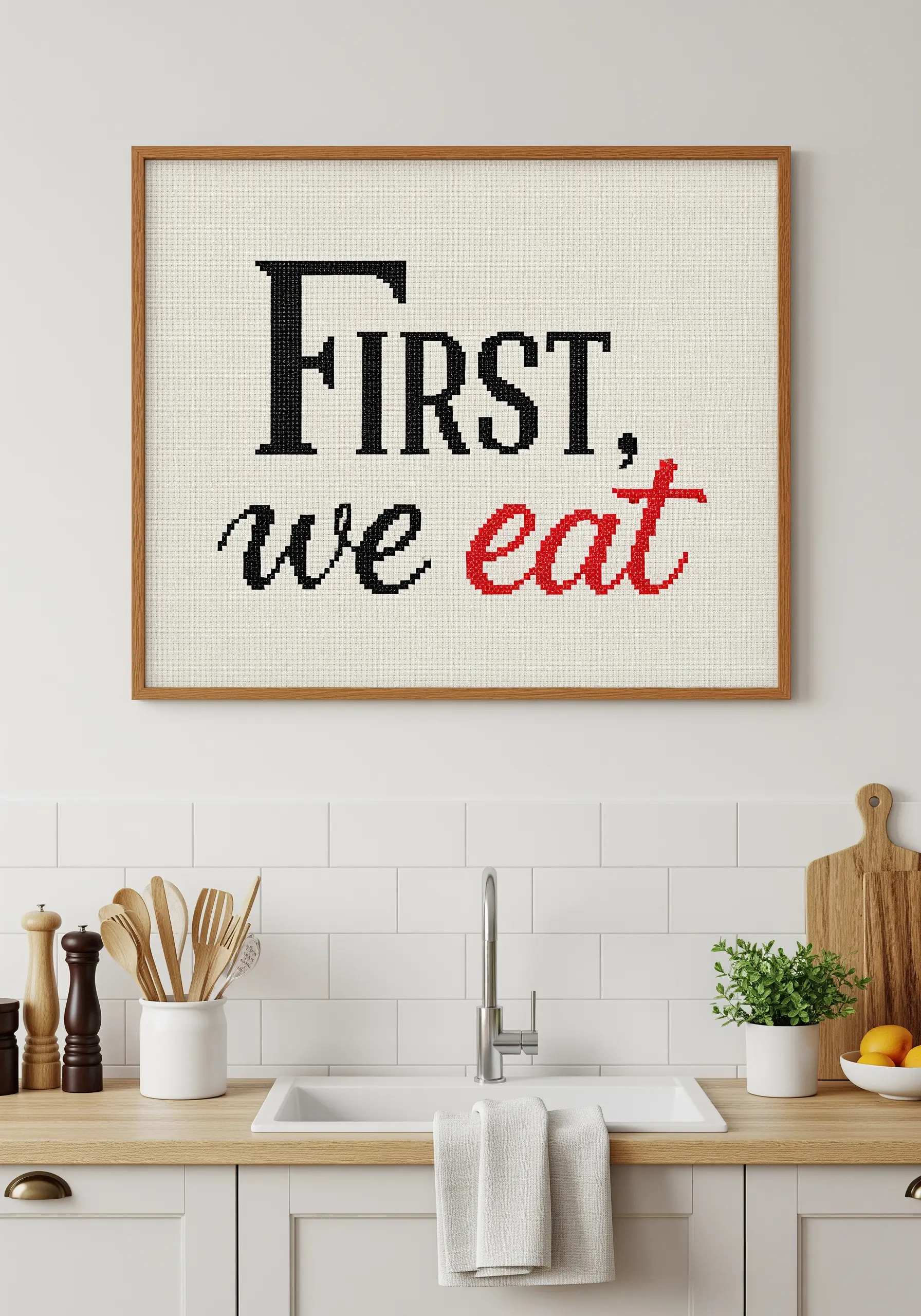 A framed cross-stitch sign for a kitchen that reads 'First, we eat' in mixed fonts.