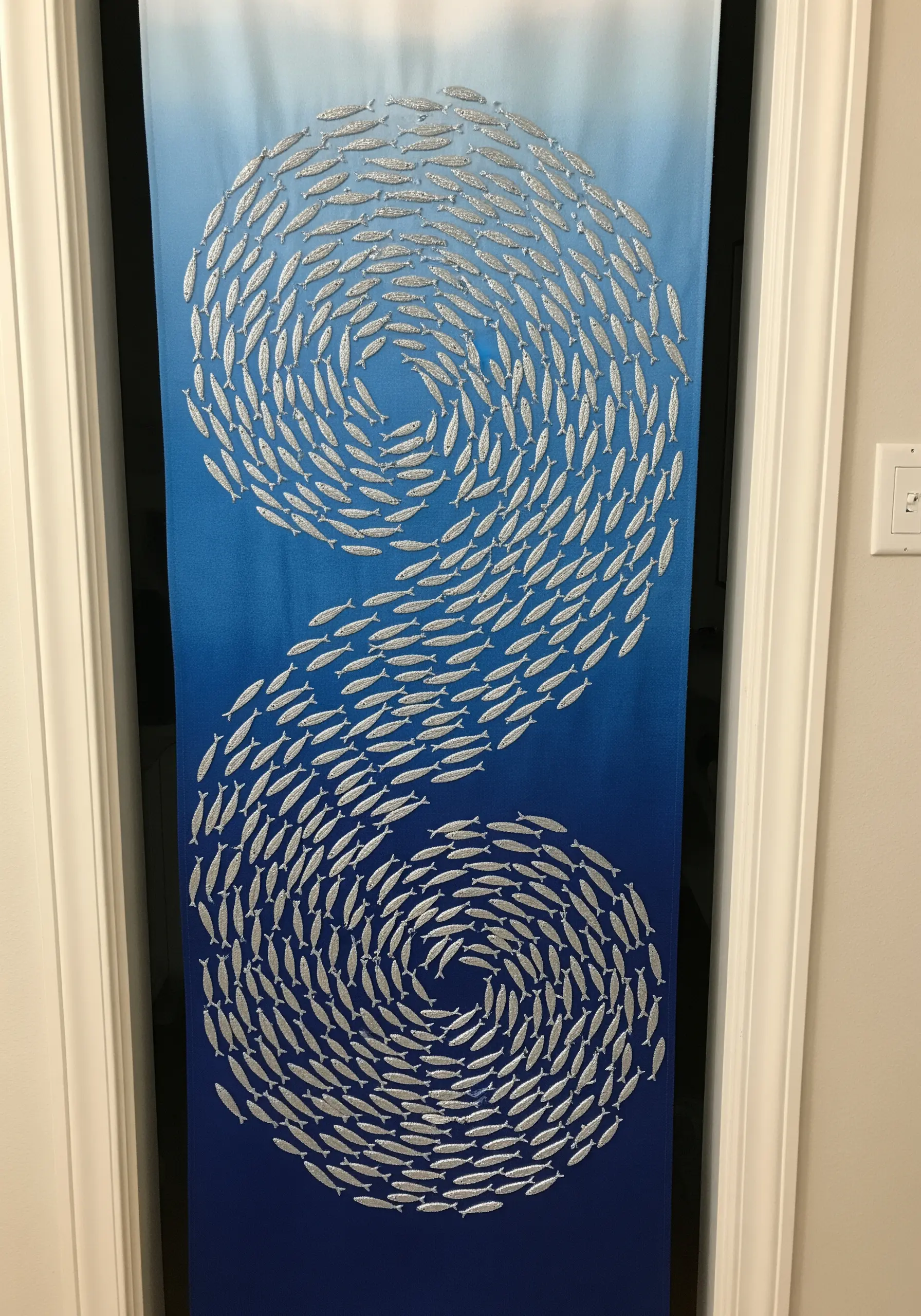 Embroidery of a swirling school of silver fish on a blue gradient fabric panel on a door.