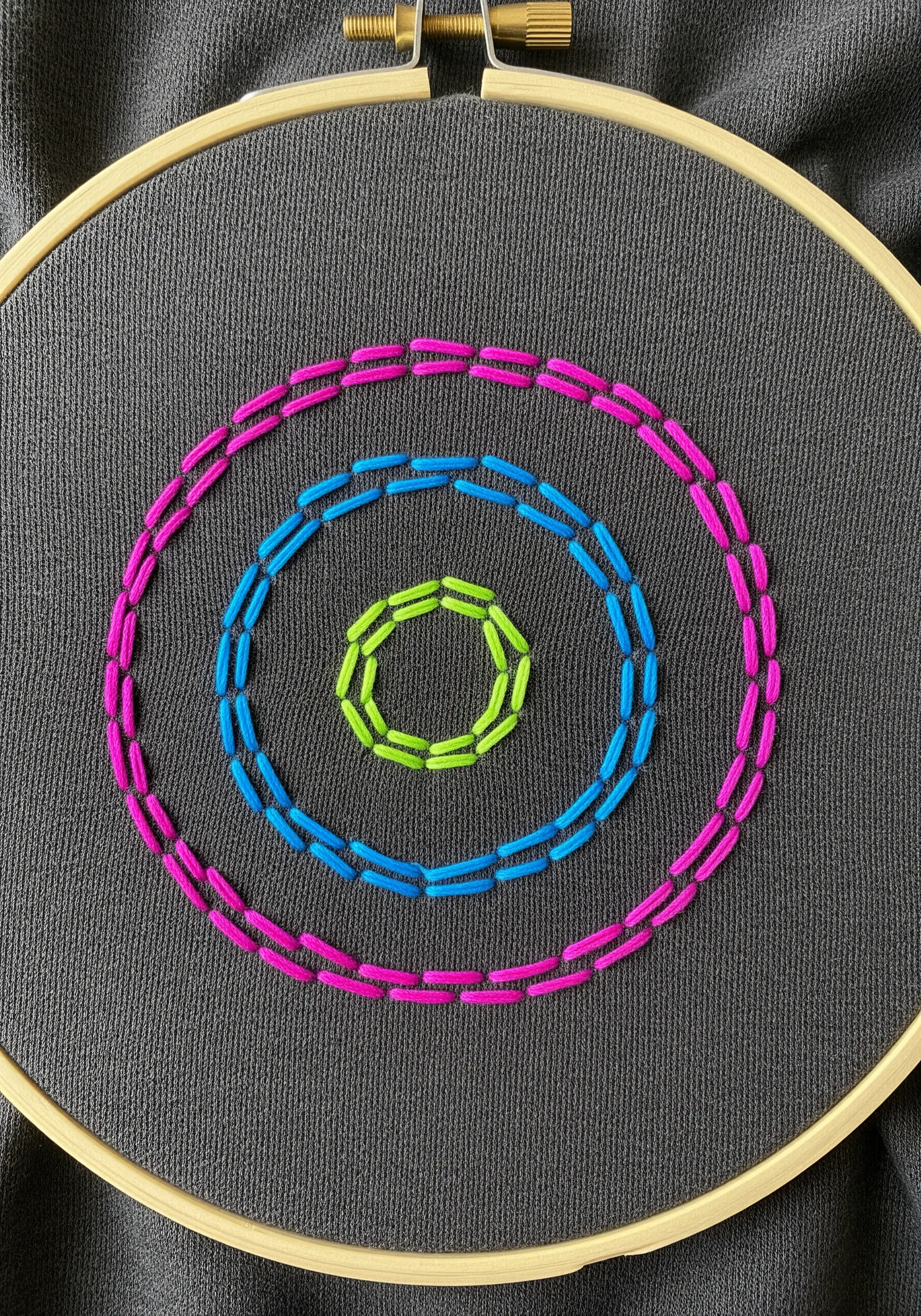 Concentric circles in neon pink, blue, and green using different stitches on dark fabric.