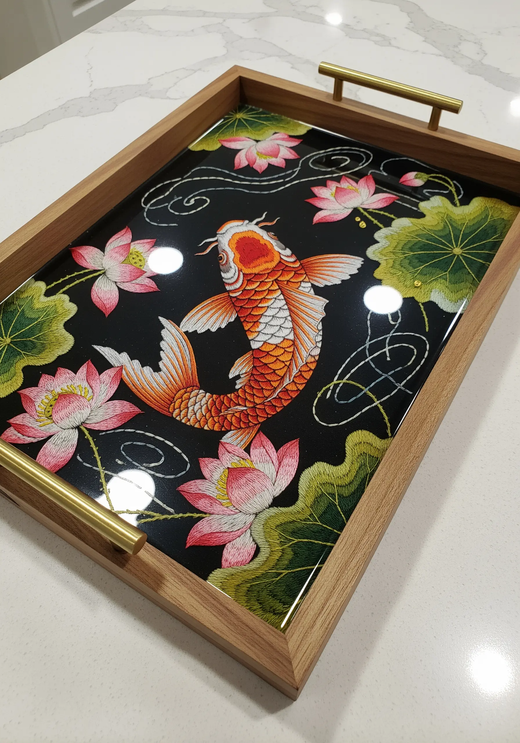 An embroidered koi fish and lotus flowers set under a clear, glossy resin in a wooden tray.