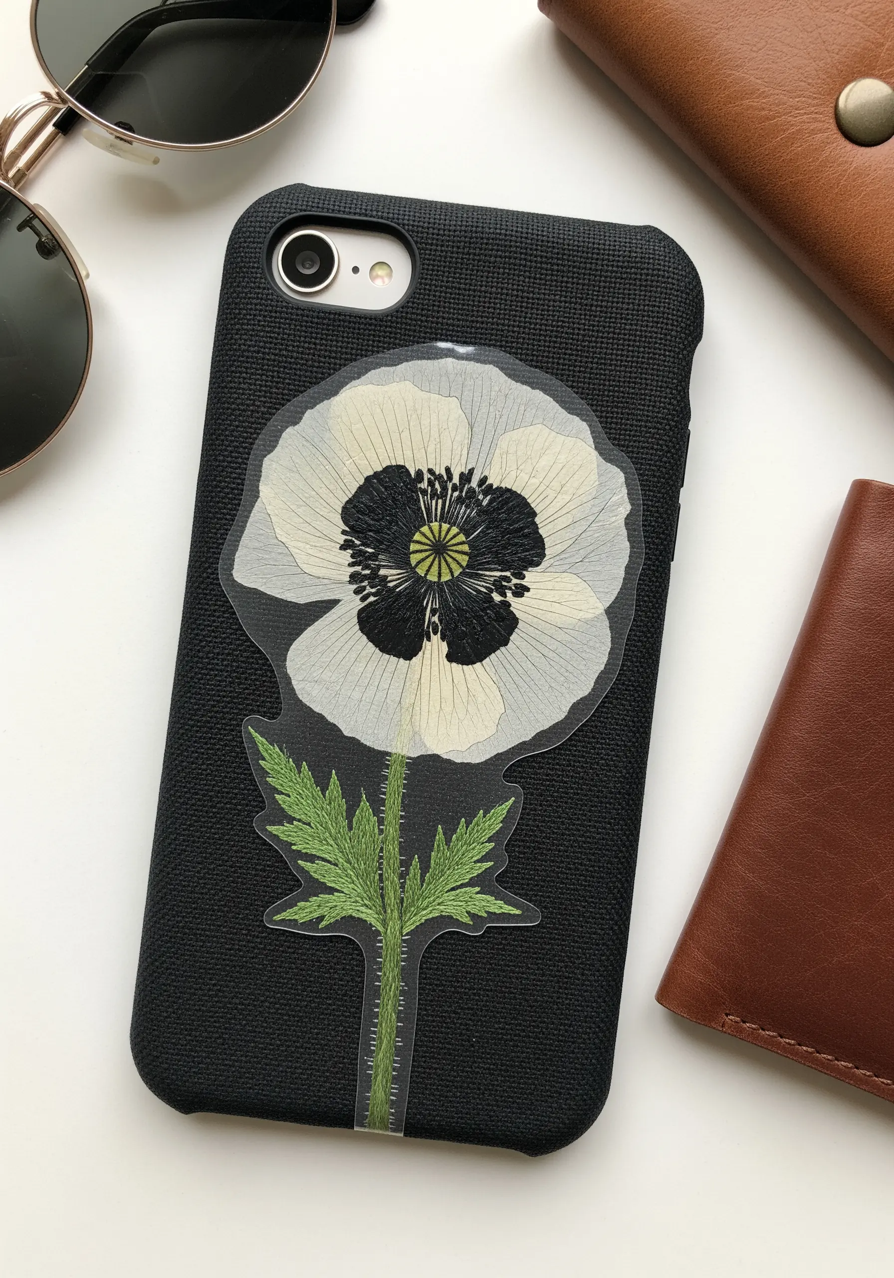 A black phone case featuring a large pressed white poppy with an embroidered stem.