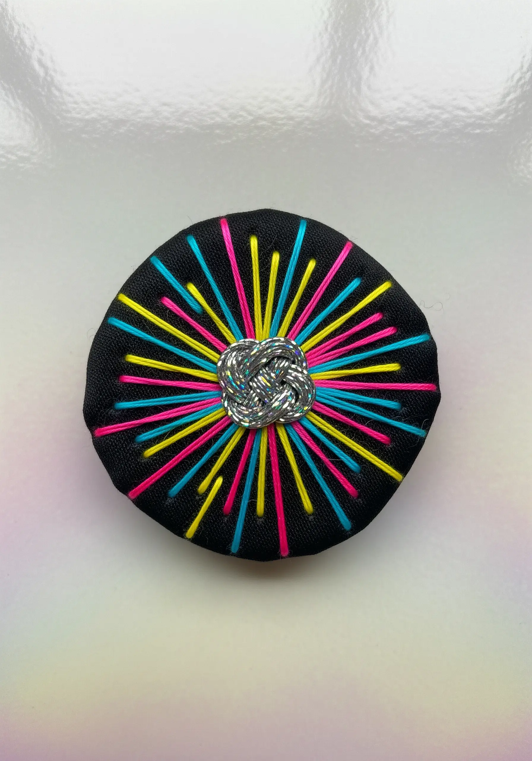 Neon straight stitches radiating from a central silver metallic knot on black fabric.