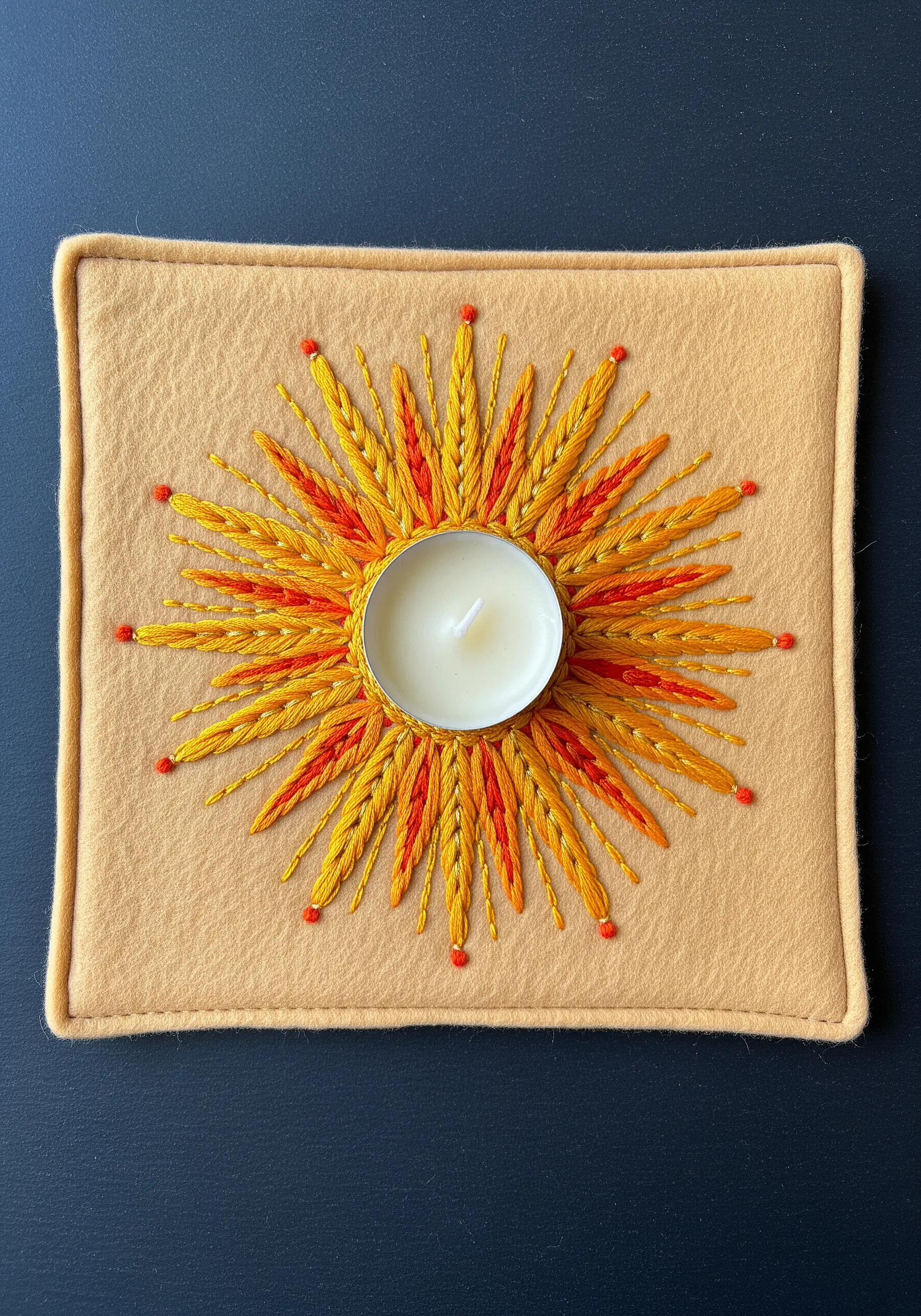 A sunburst motif embroidered in yellow and orange on a square felt mat holding a tealight candle.