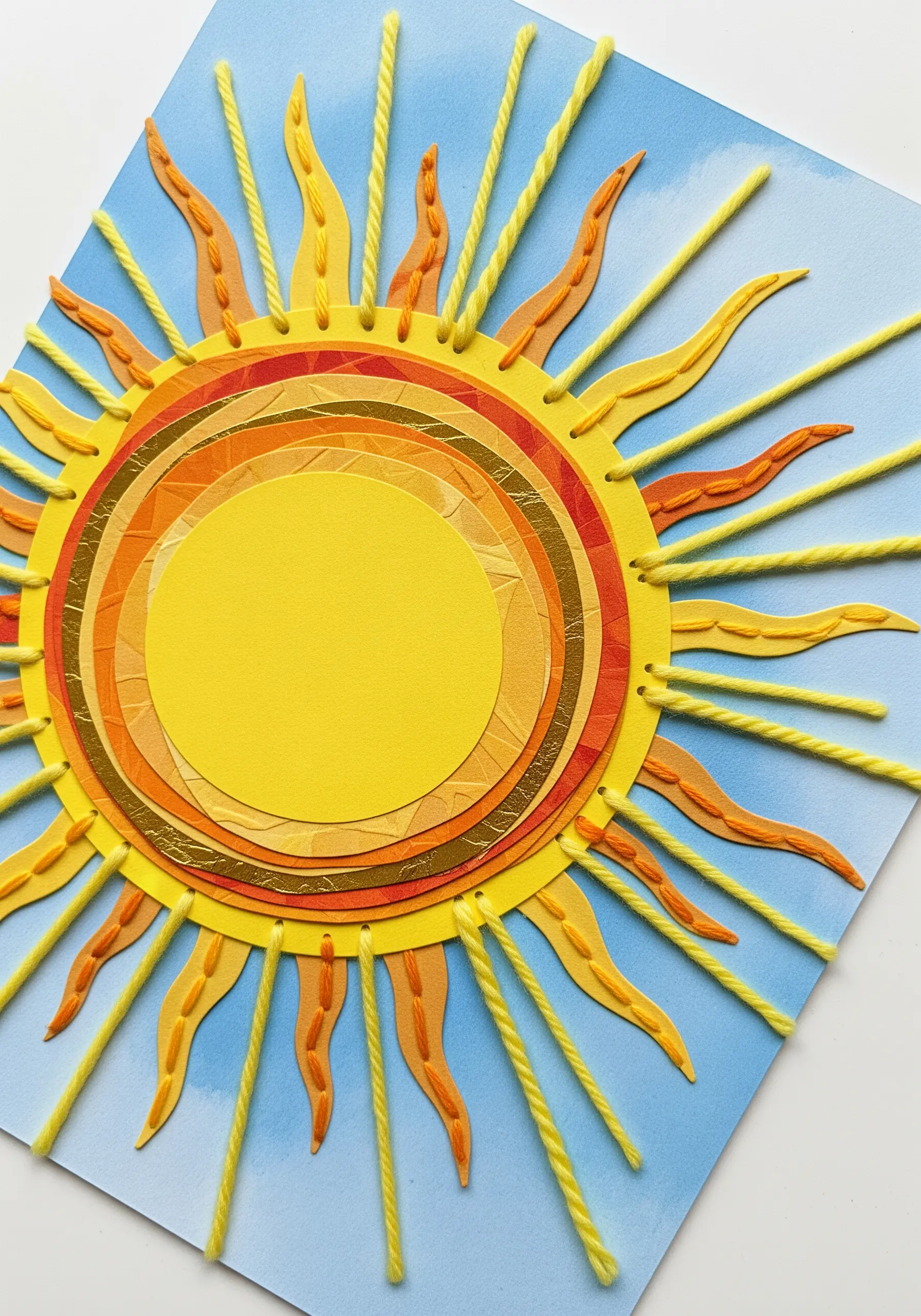 A layered paper sun with bright yellow yarn stitched in long, radiating rays.