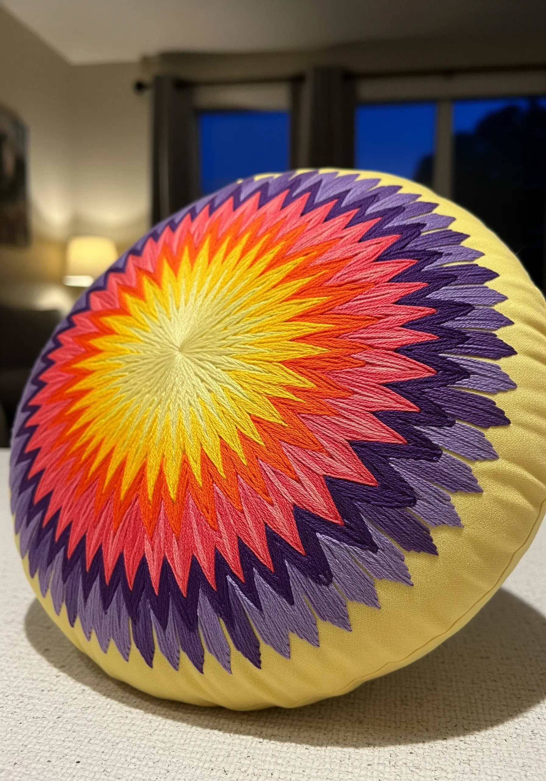 A round pillow with a radiating spike mandala embroidered in a gradient of yellow, pink, and purple.