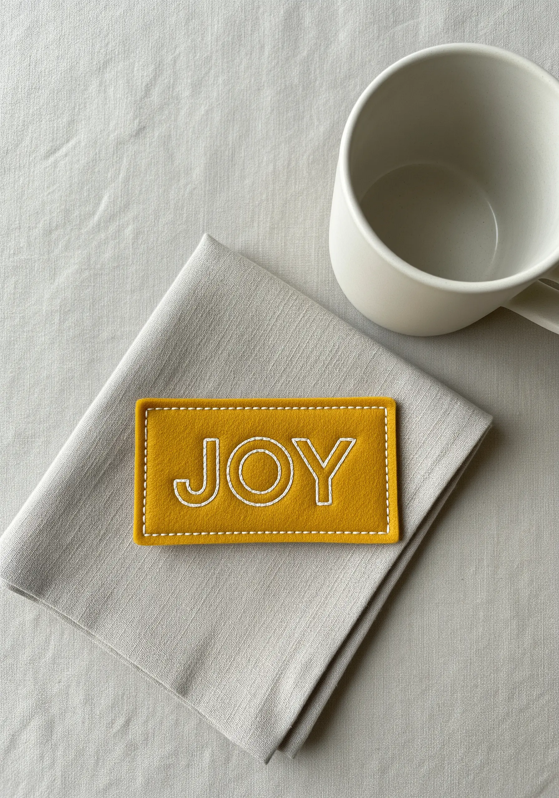 A yellow felt patch with the word 'JOY' stitched in a clean, white outline font.