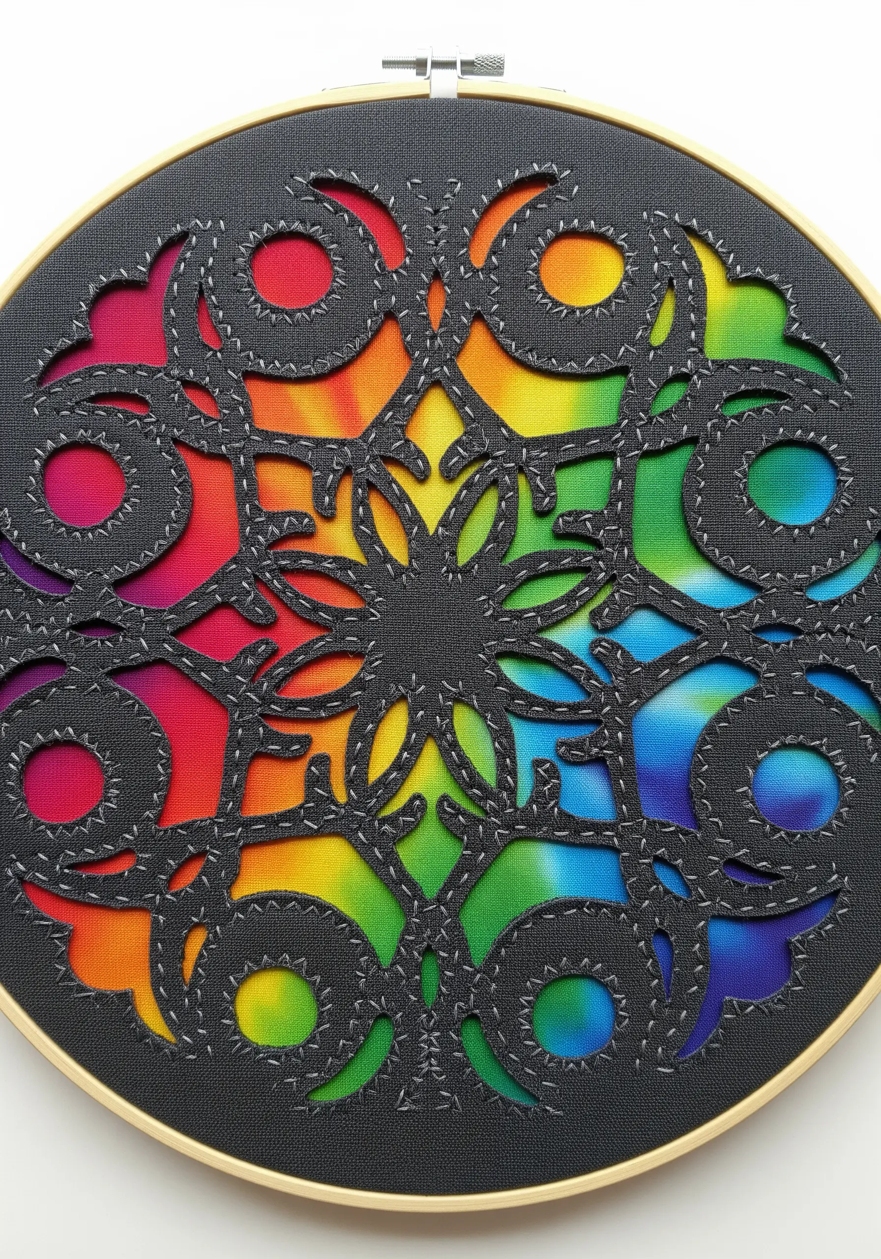 A black cutwork mandala laid over a vibrant rainbow tie-dye fabric, stitched in a hoop.