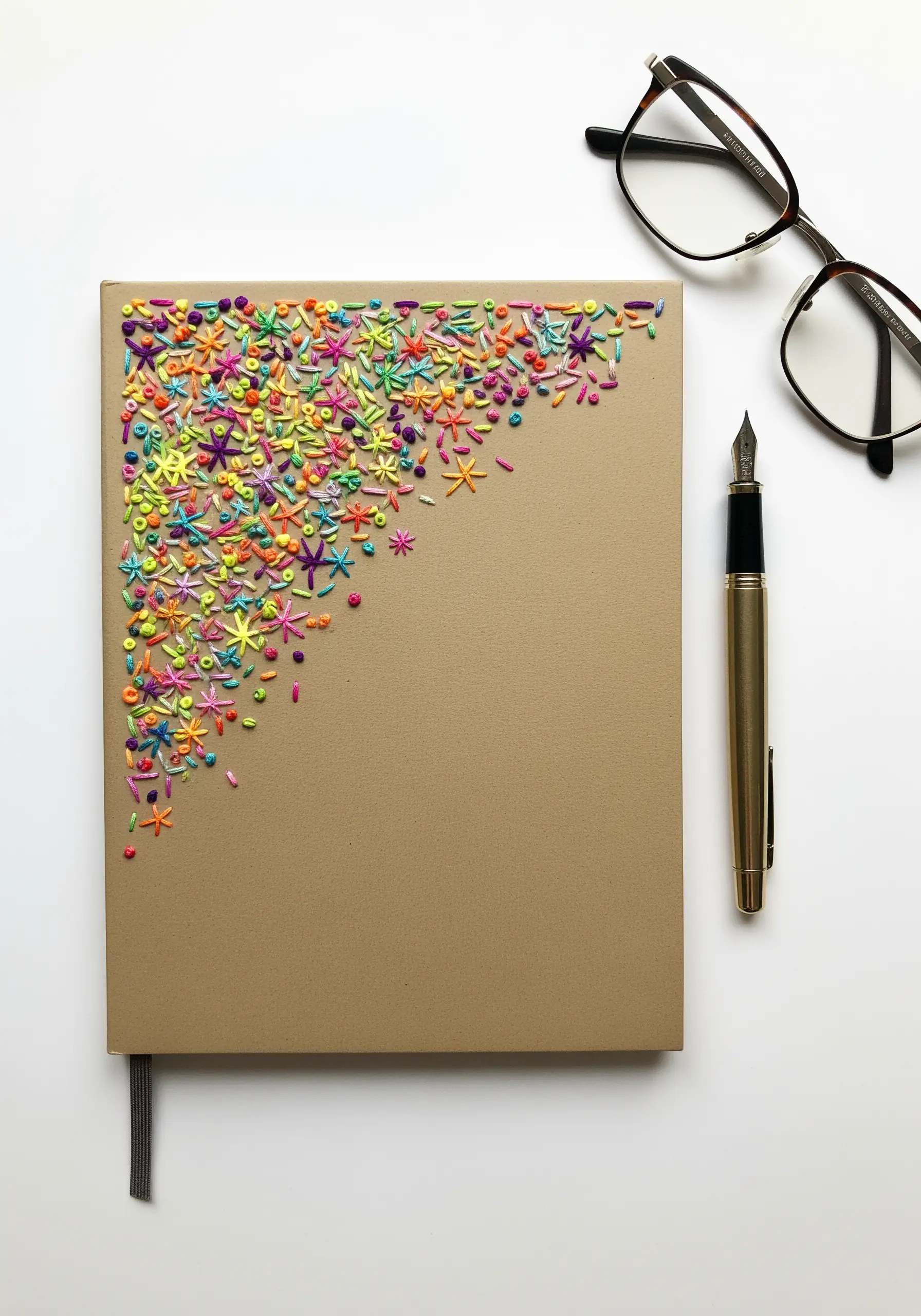 The corner of a kraft paper journal covered in a dense confetti of tiny, colorful embroidery stitches.