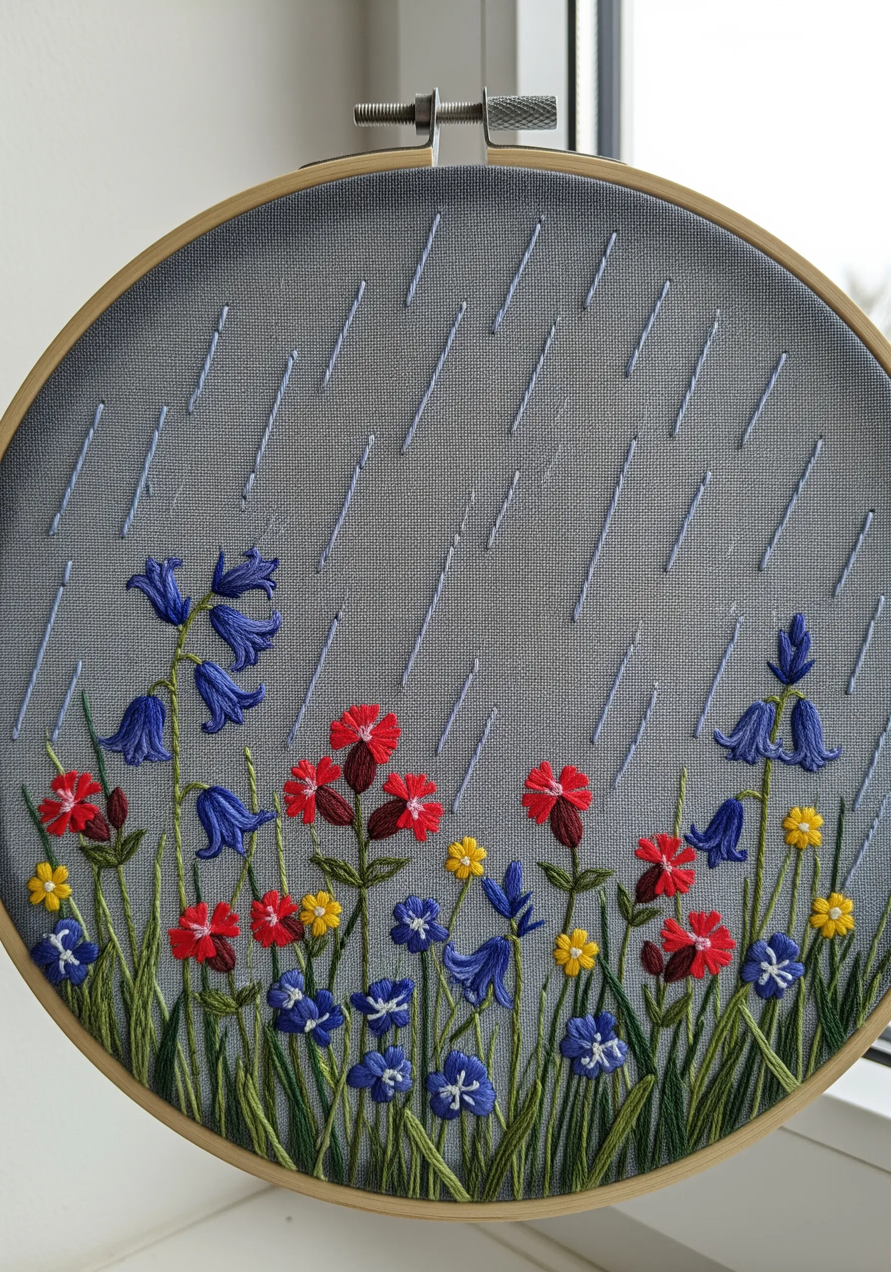 Embroidery of wildflowers under diagonal stitches representing falling rain on a grey background.
