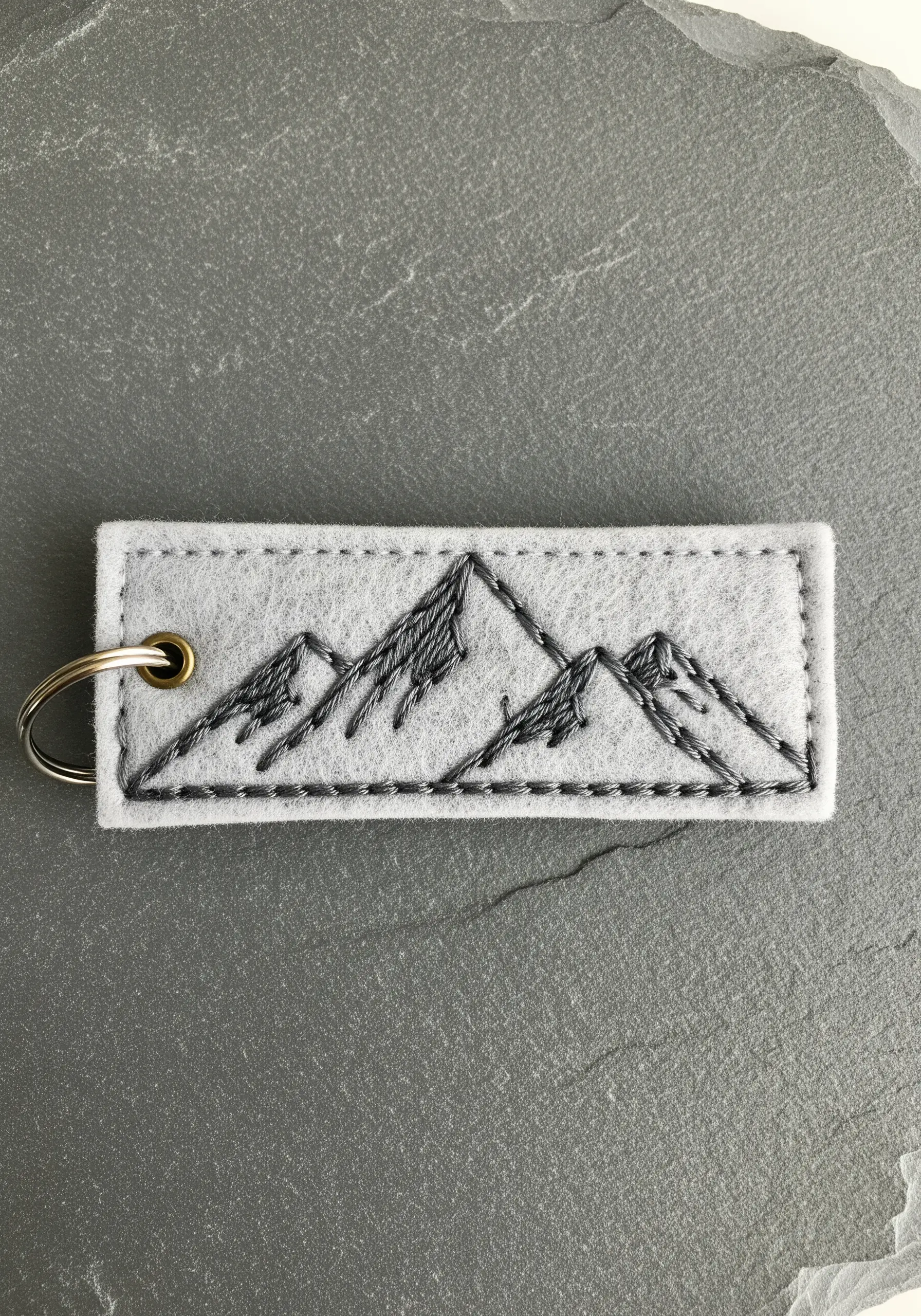 Black and gray sketch-style embroidery of mountains on a gray felt keychain