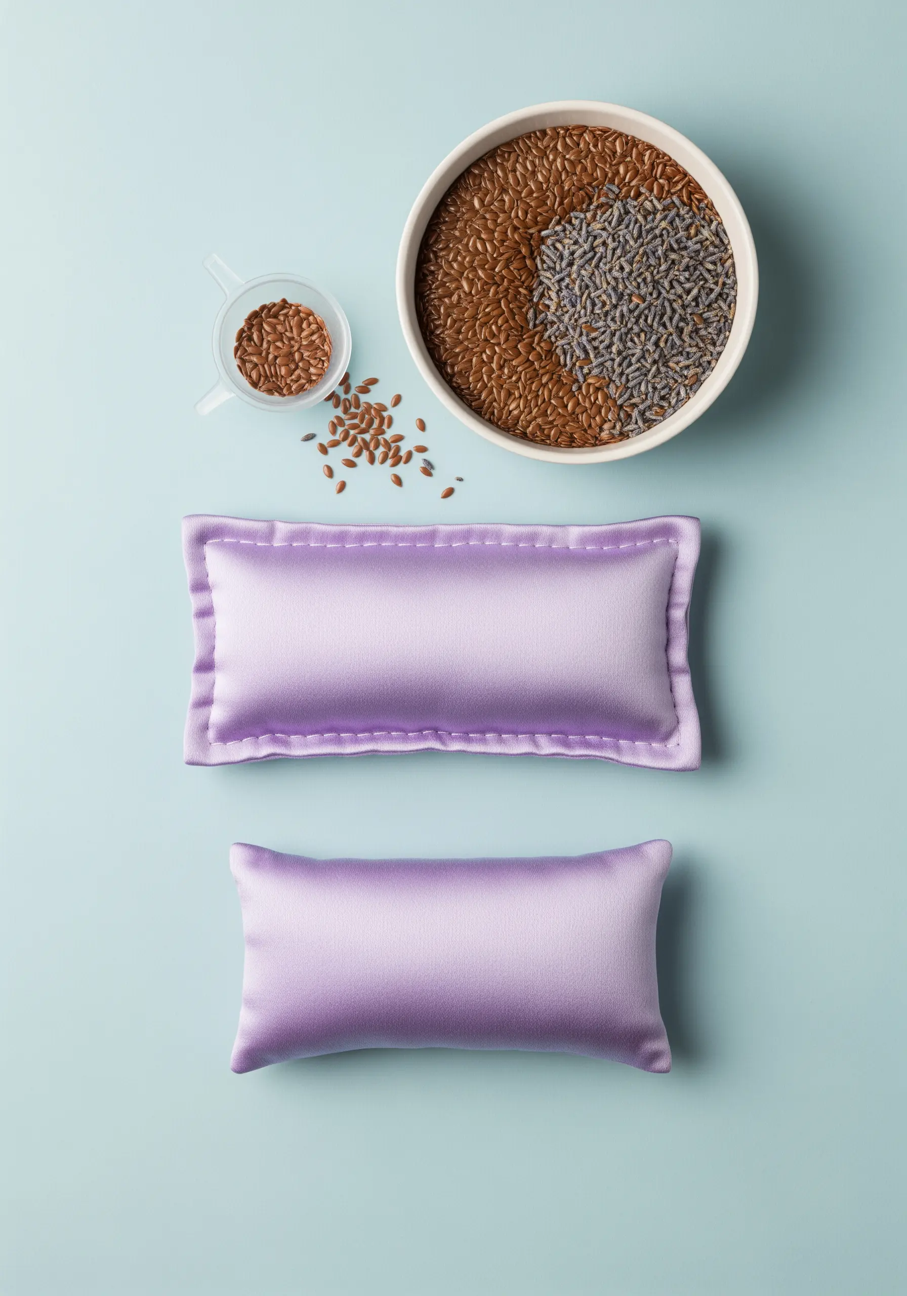 Two lavender-colored satin eye pillows with a bowl of flax seeds and lavender buds.