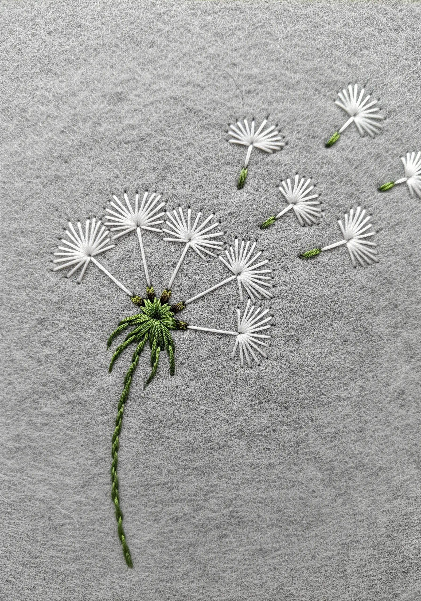 A dandelion puff with seeds blowing away, stitched in white and green on gray felt.