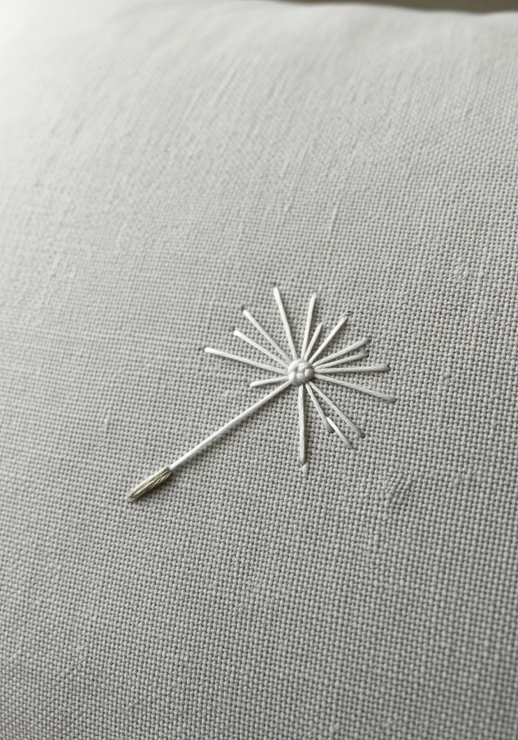 A single dandelion seed embroidered in white thread on a light gray linen pillowcase.