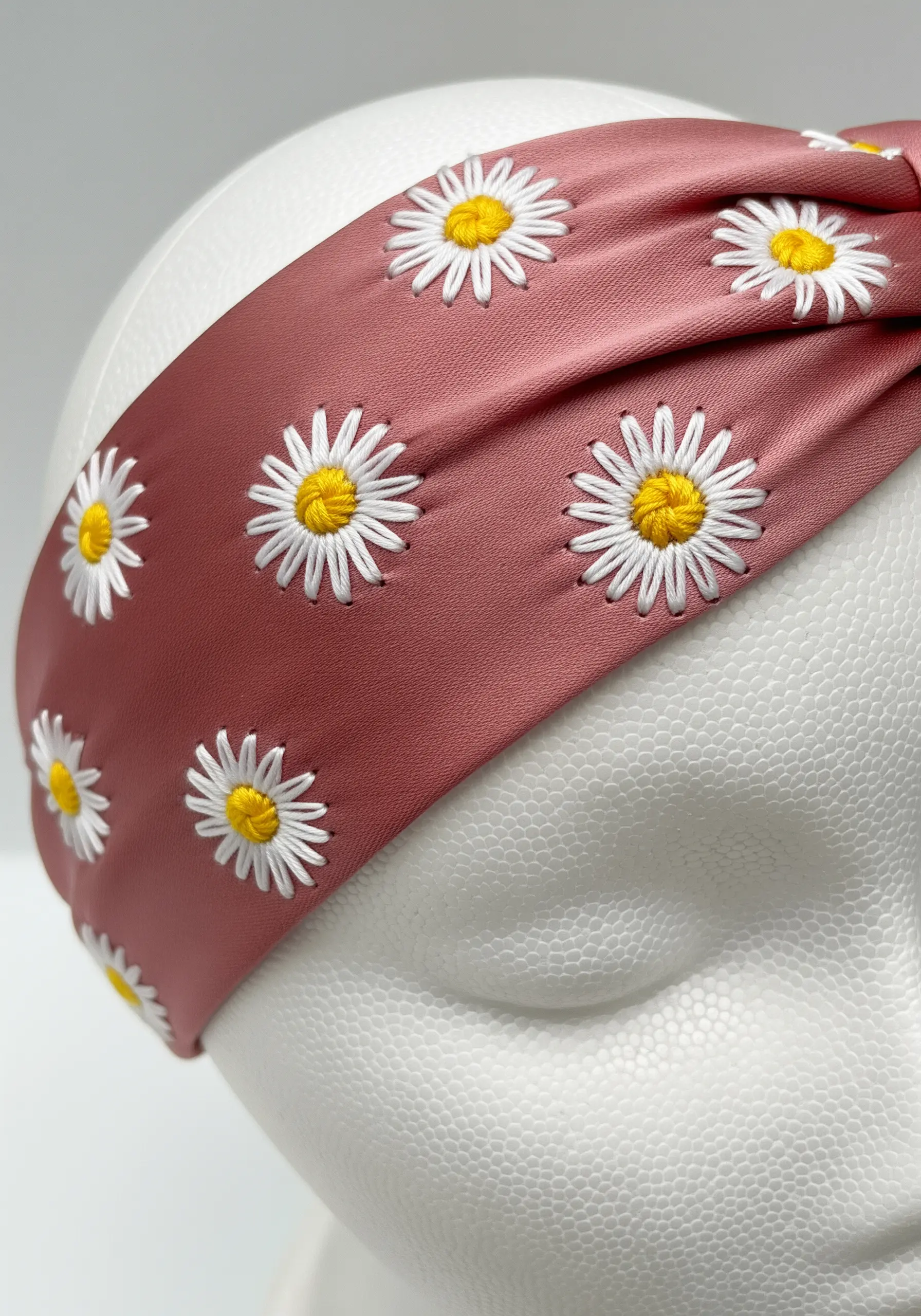 A dusty rose fabric headband adorned with a repeating pattern of embroidered daisies.