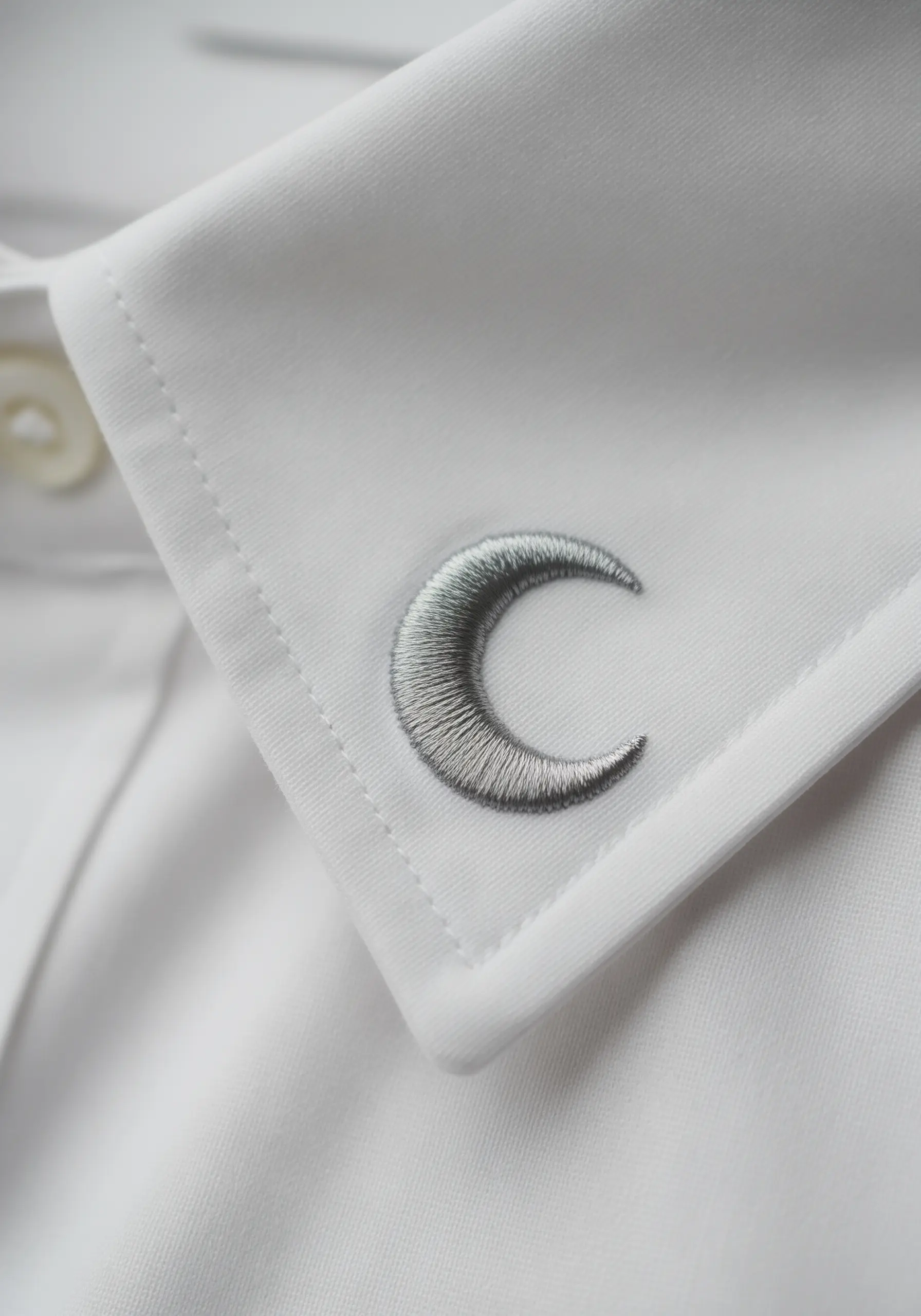 A small, realistic crescent moon with a gray gradient, embroidered on a white shirt cuff.