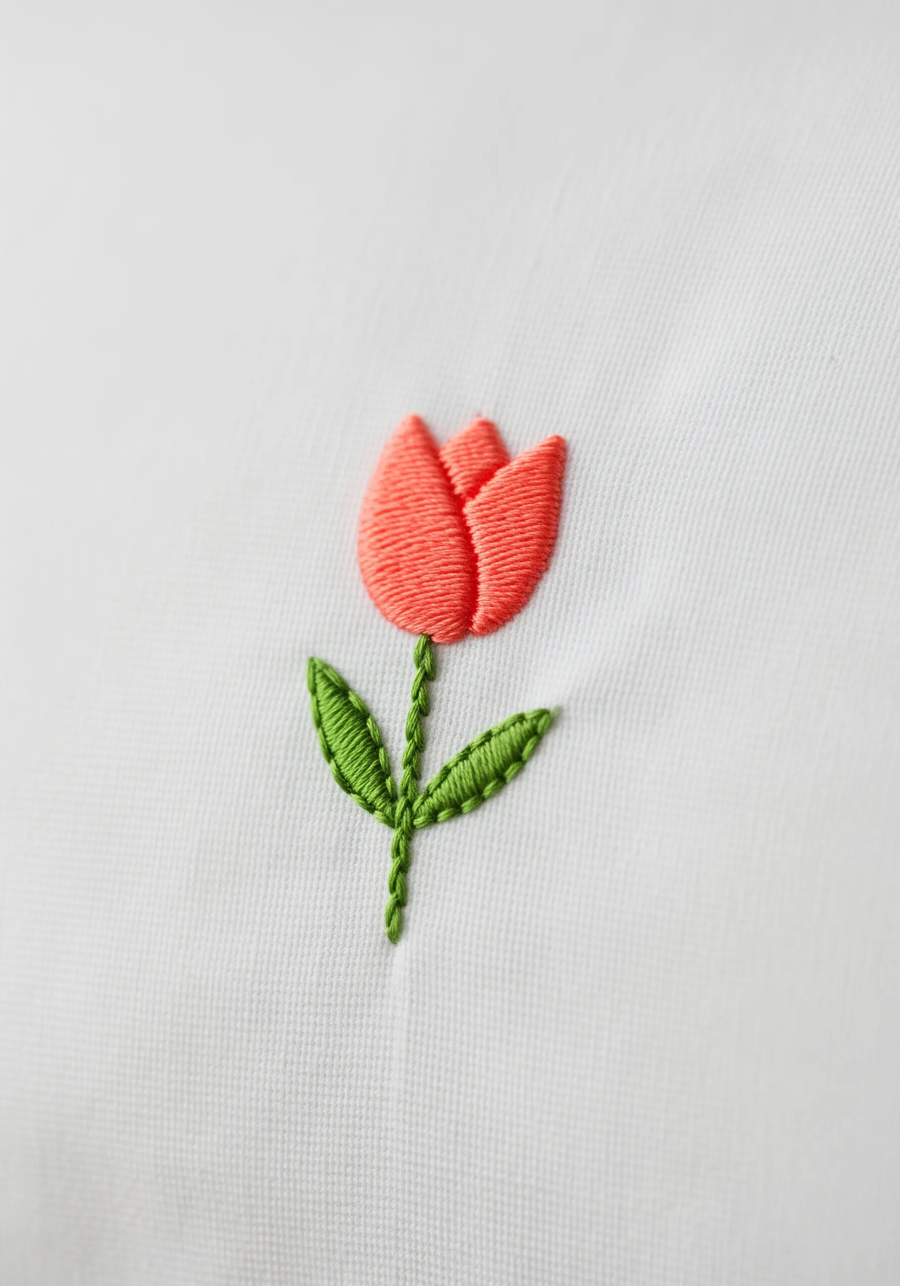 A small, simple tulip with coral petals and green leaves embroidered on a white pillowcase.