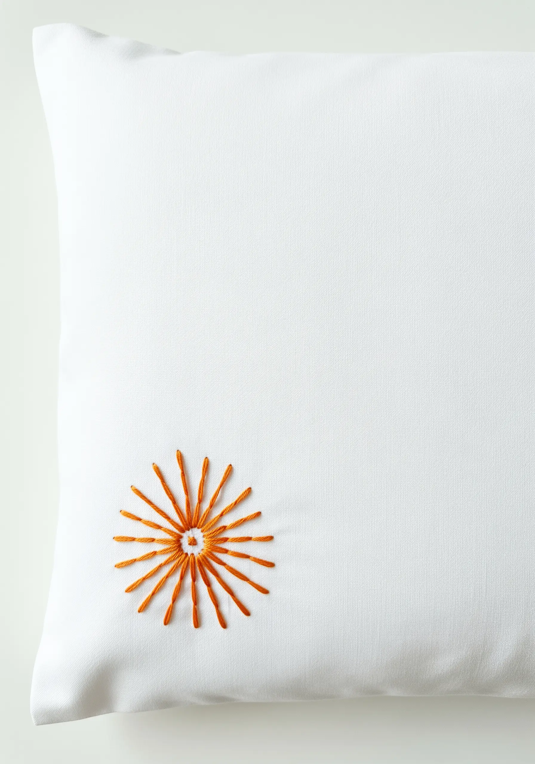 A stylized sunburst embroidered in orange thread on the corner of a white pillowcase.