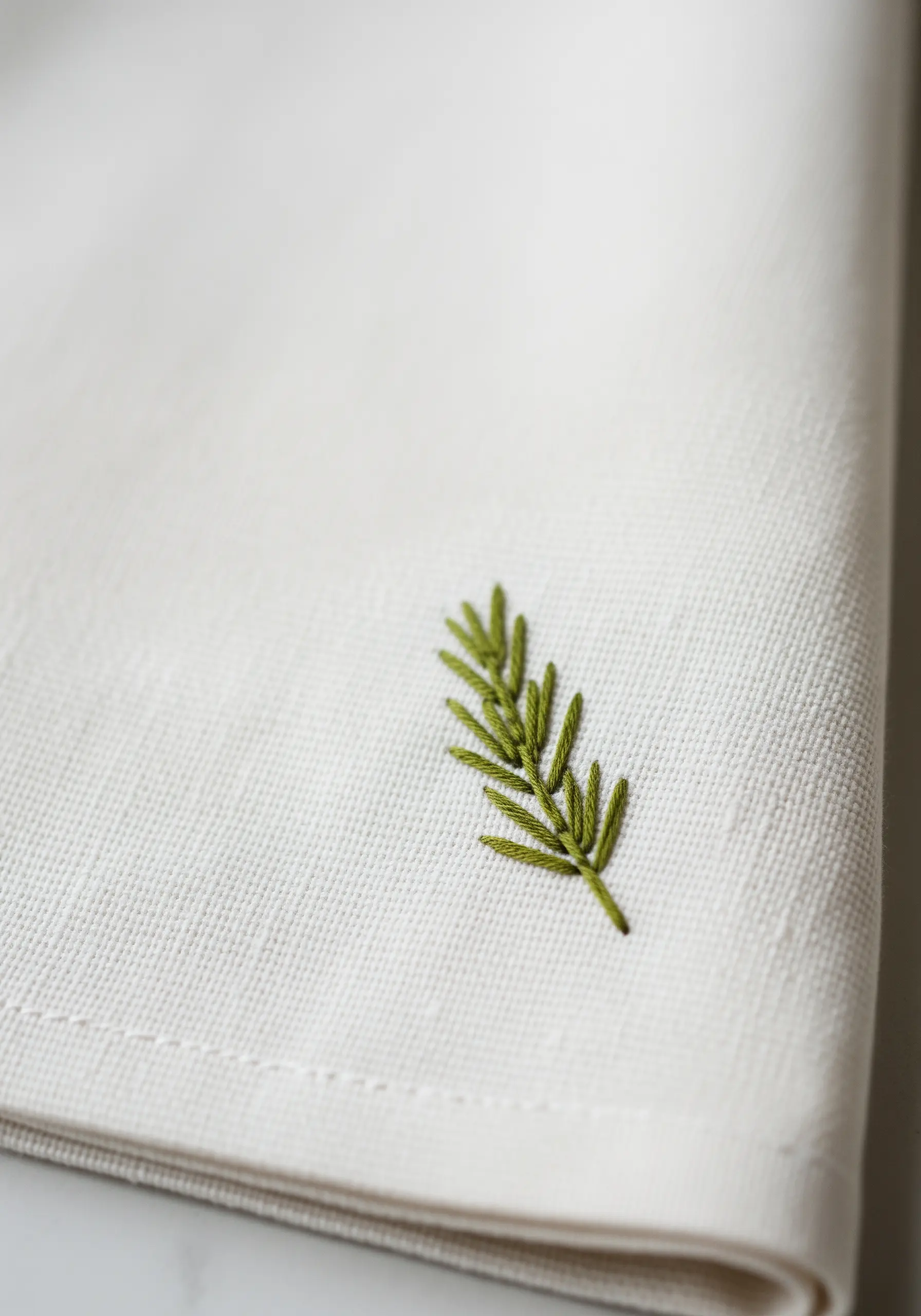 A single sprig of rosemary embroidered on the corner of an off-white linen napkin.