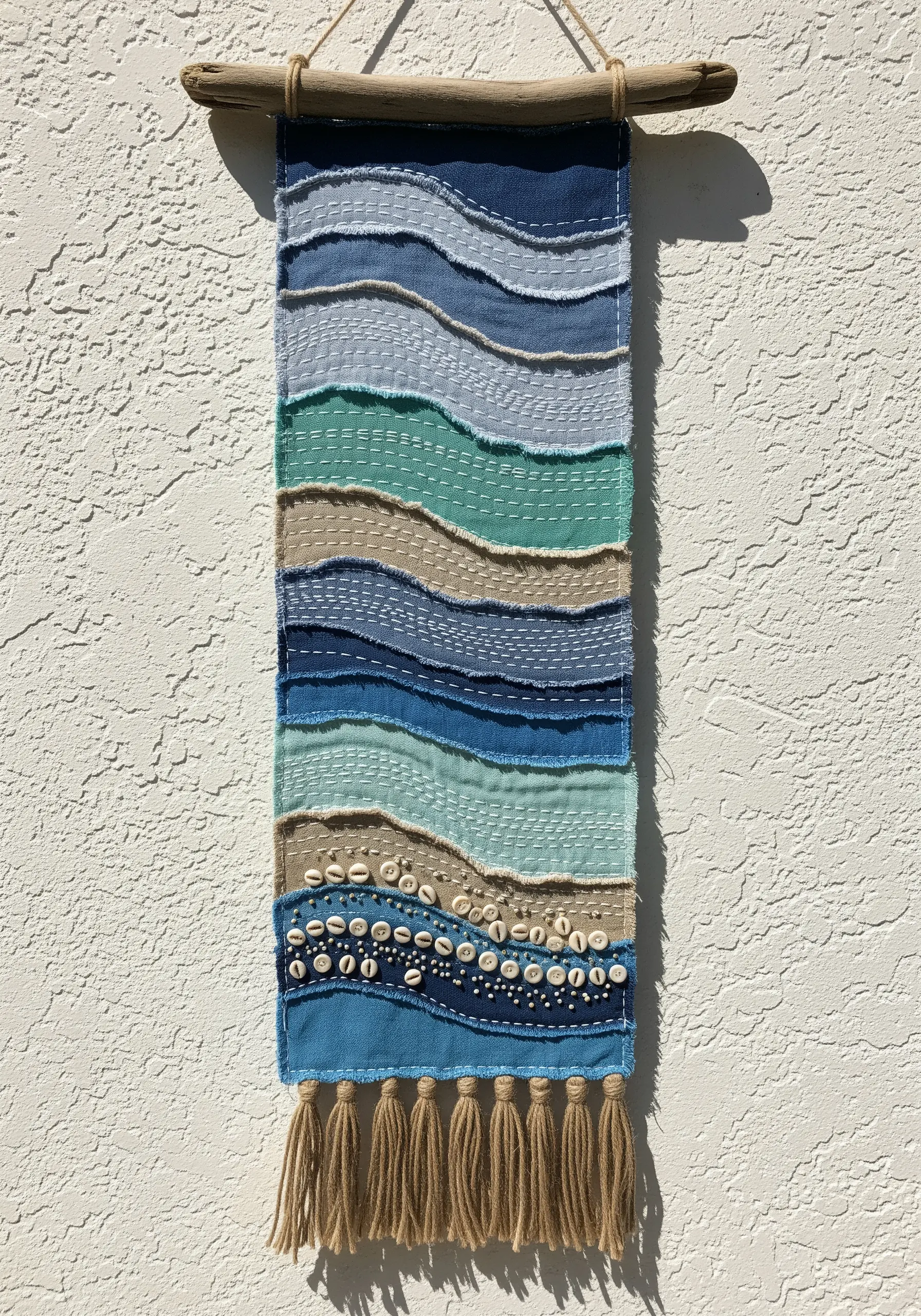 Coastal wall hanging made of layered blue fabric strips with running stitches and shells.