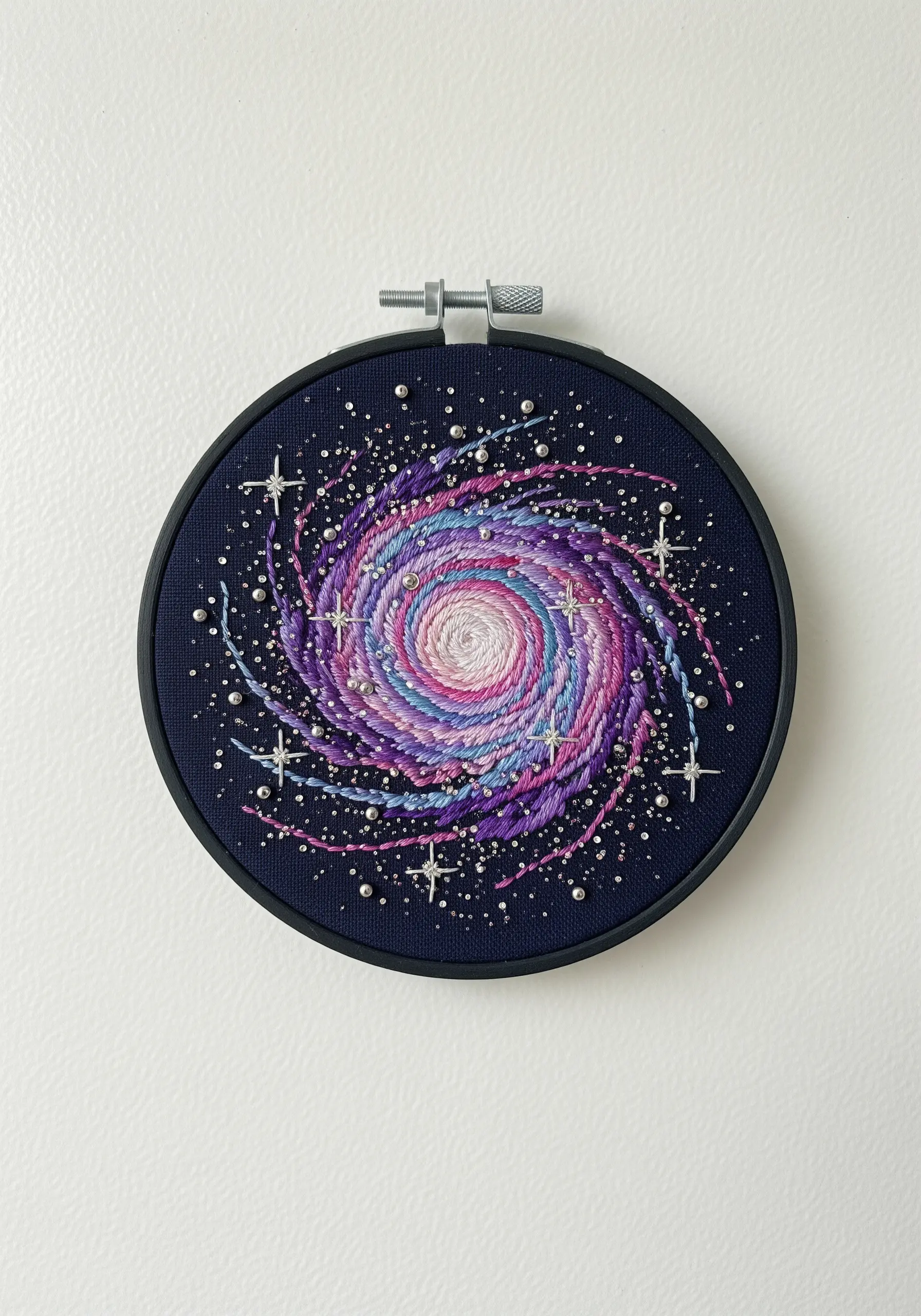 An embroidered spiral galaxy in pink, purple, and blue on dark fabric in a black hoop.