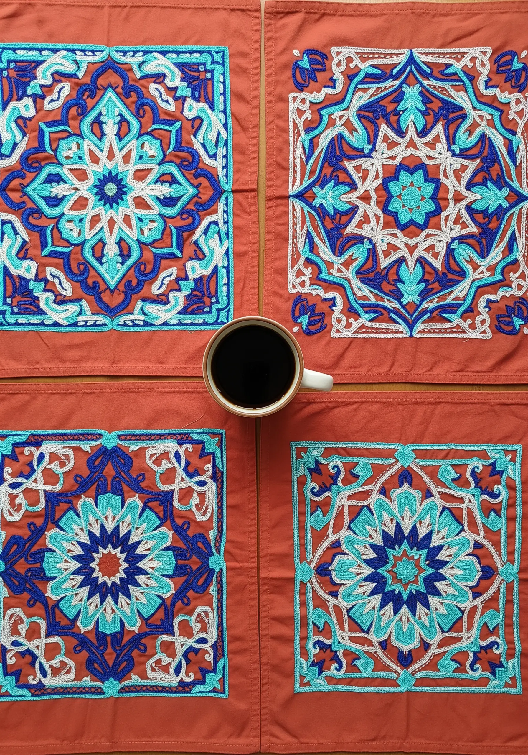 Four terracotta-colored placemats, each with a detailed embroidered mosaic pattern in blue and white.