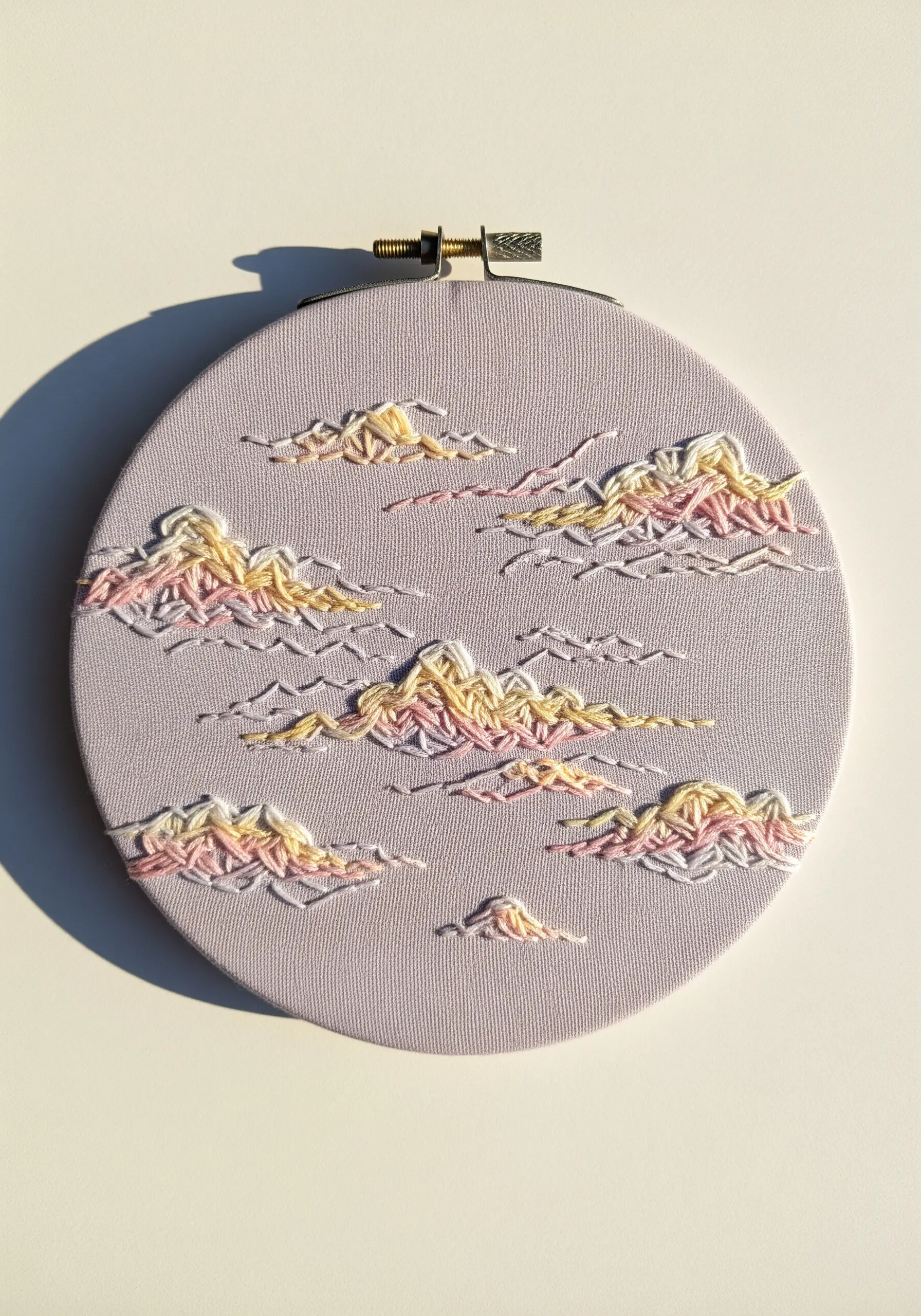 Embroidery hoop with soft, wispy clouds stitched in pastel pink and yellow on lavender fabric.