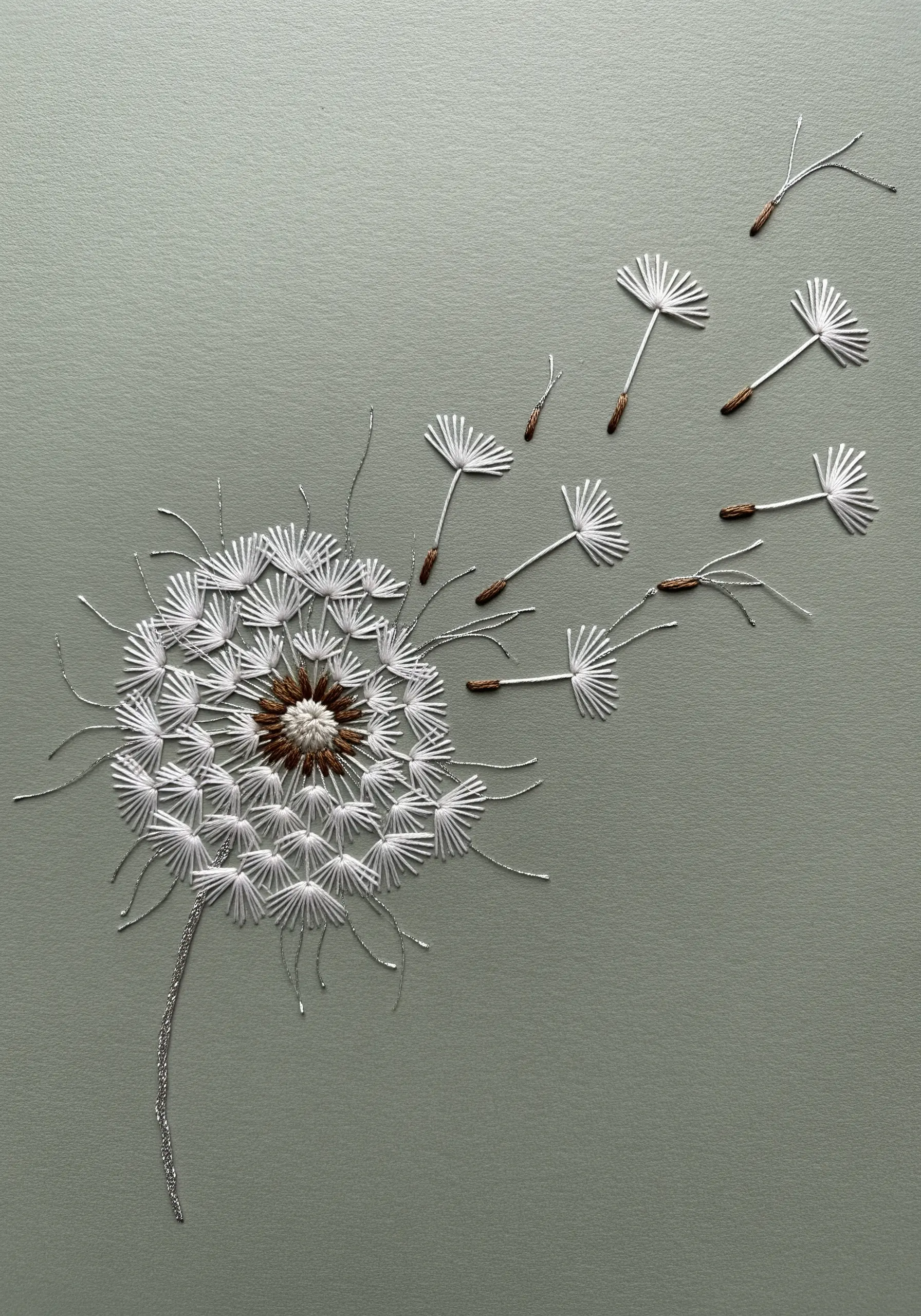 A dandelion puff ball embroidered with delicate white straight stitches and metallic thread on gray paper.