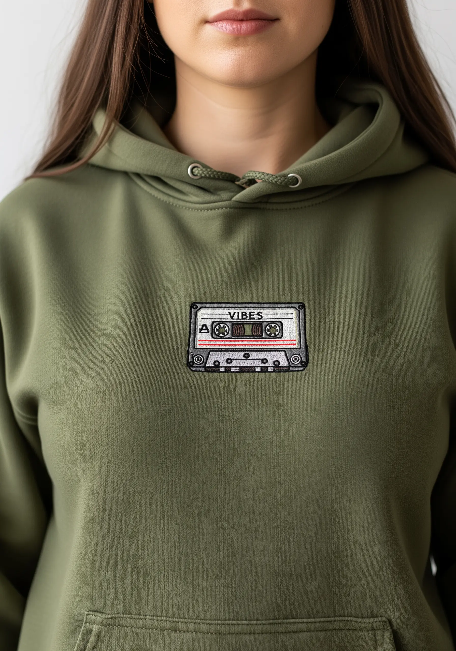 A small, detailed embroidered patch of a retro cassette tape on an olive green hoodie.
