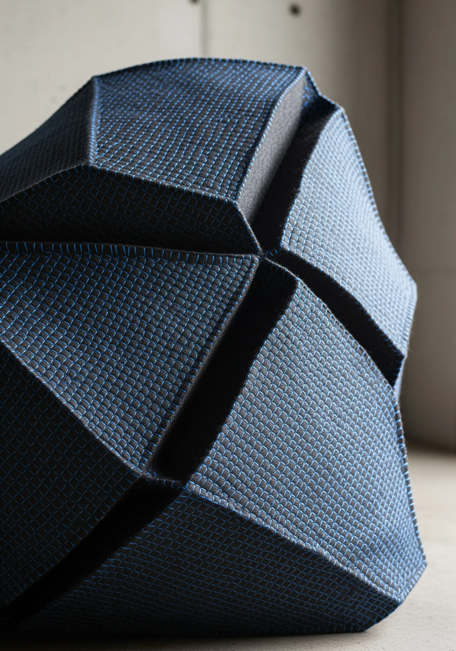 A geometric, faceted 3D object covered in dark blue fabric with a light blue grid stitch pattern.