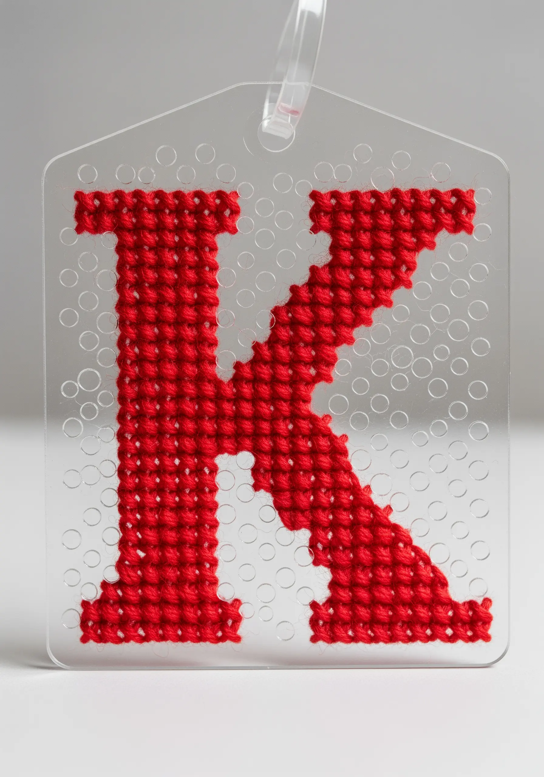 A bright red cross-stitched letter 'K' on a clear plastic canvas tag.