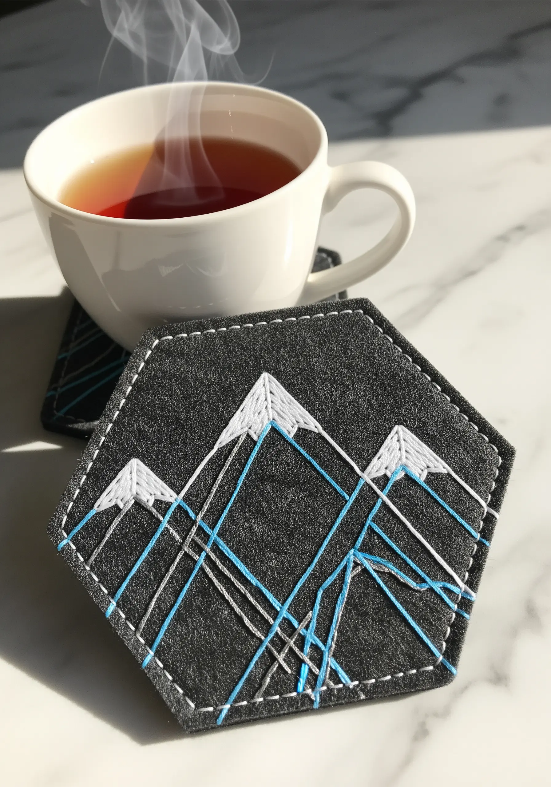 A hexagonal felt coaster with a minimalist geometric mountain range stitched in white and blue.