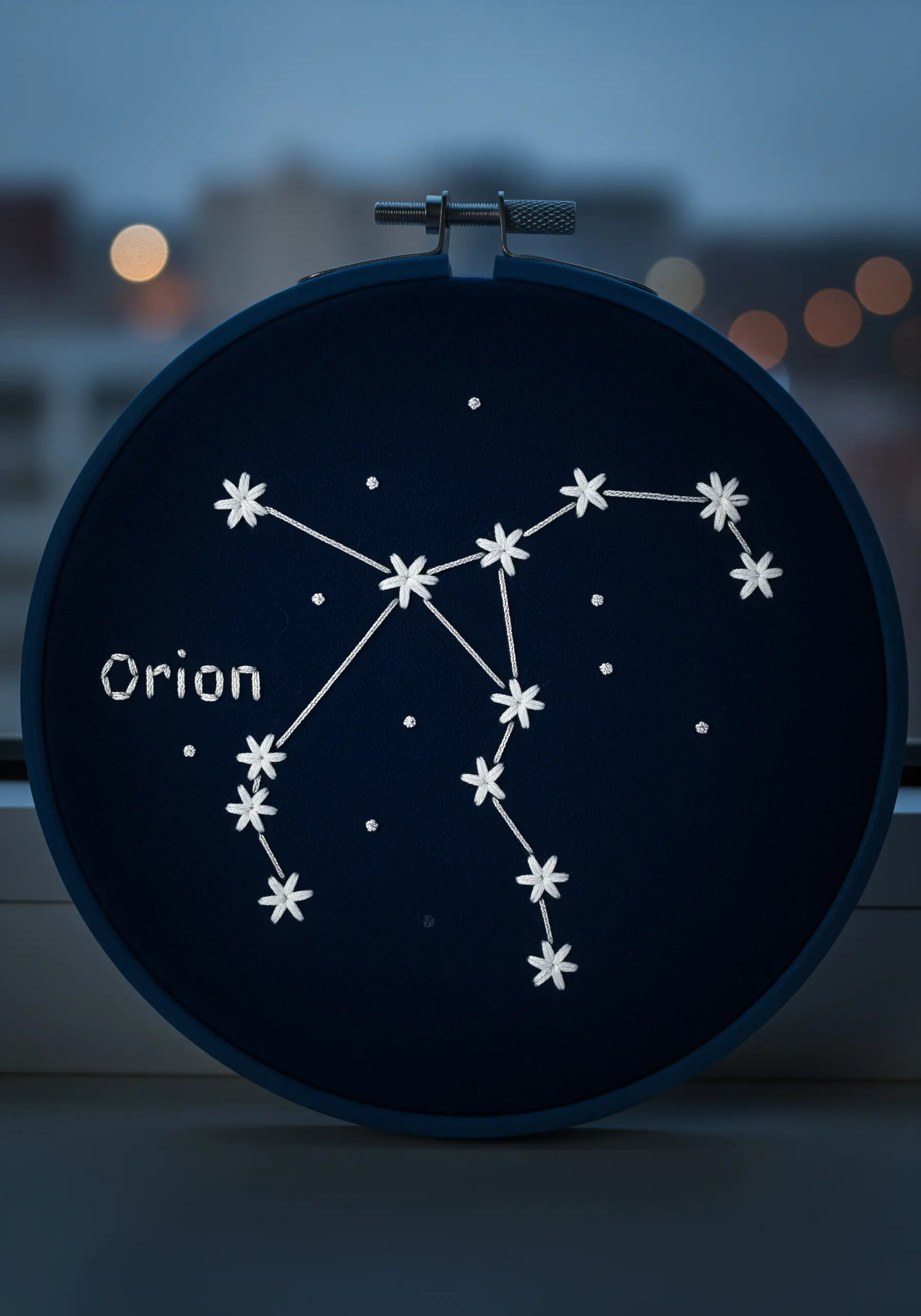 An embroidery of the Orion constellation stitched in white thread on dark navy fabric in a hoop.