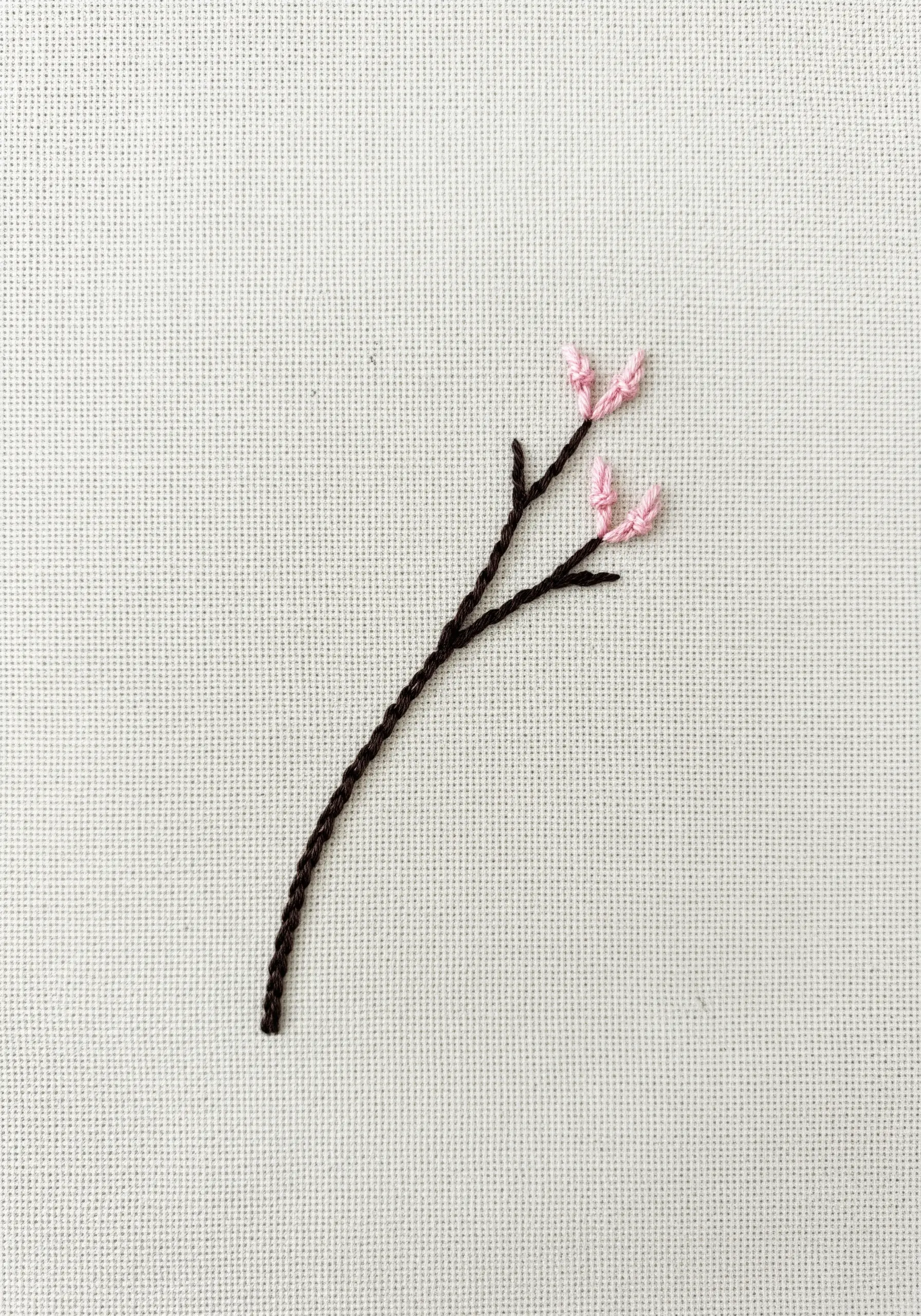 Minimalist embroidery of a dark brown branch with three tiny pink blossoms.