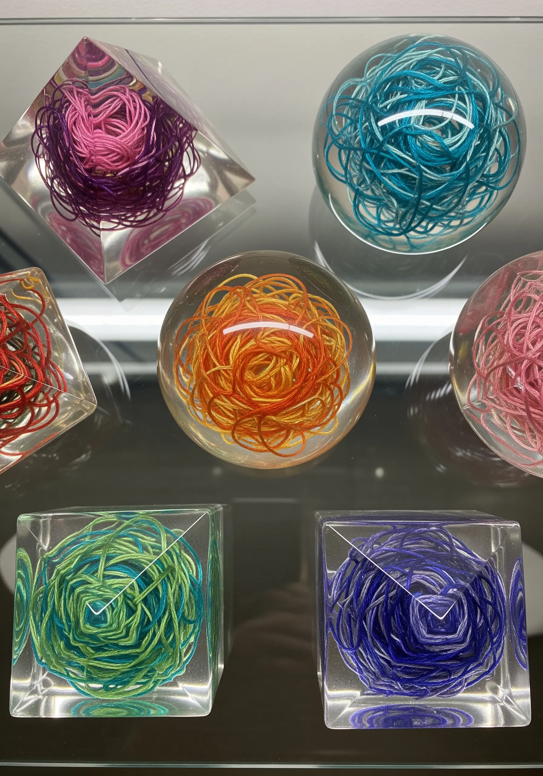 Colorful tangled thread balls suspended in clear resin spheres and cubes on a glass shelf.