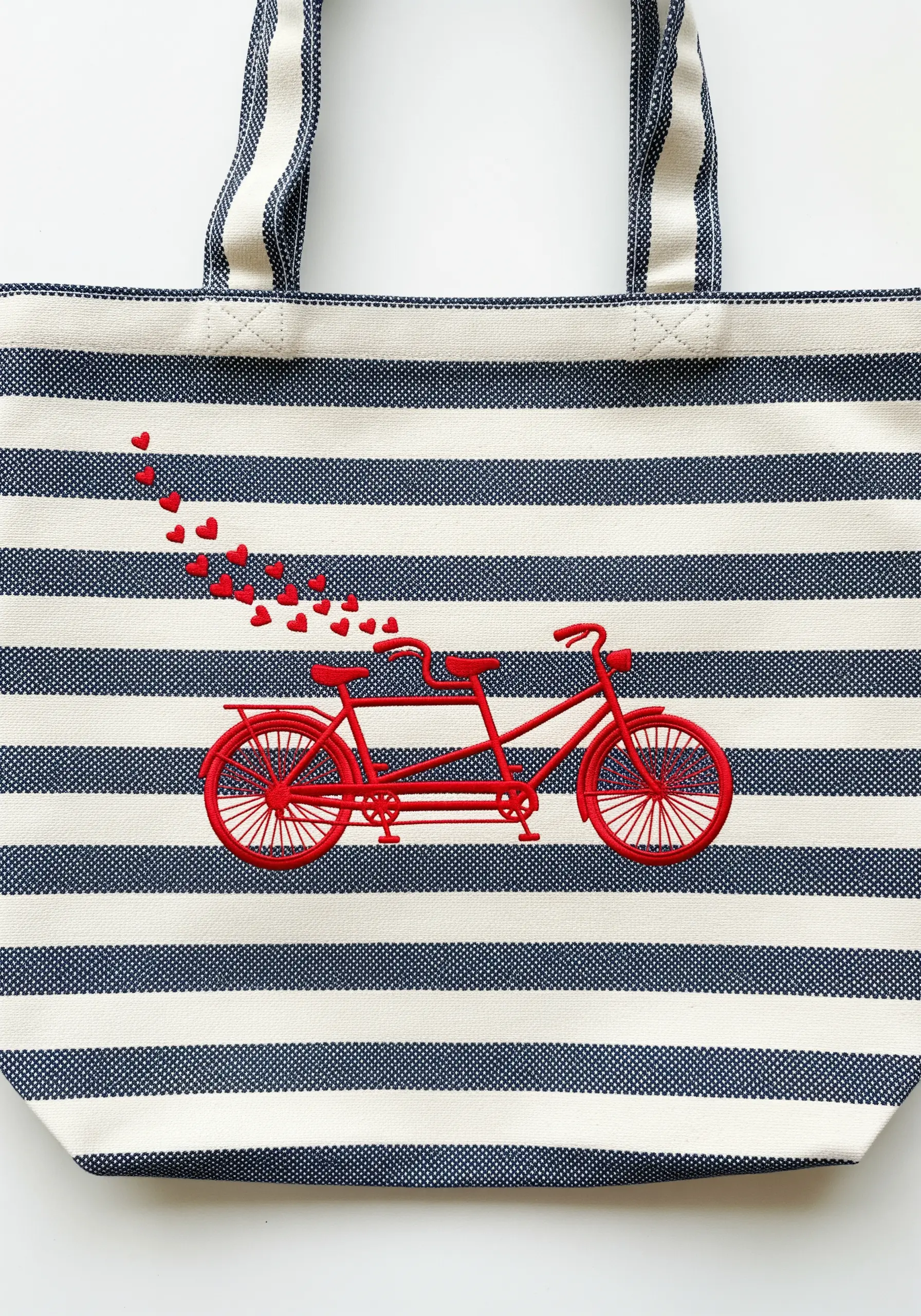 A red tandem bicycle embroidered on a blue and white striped tote bag, with tiny red hearts floating away.