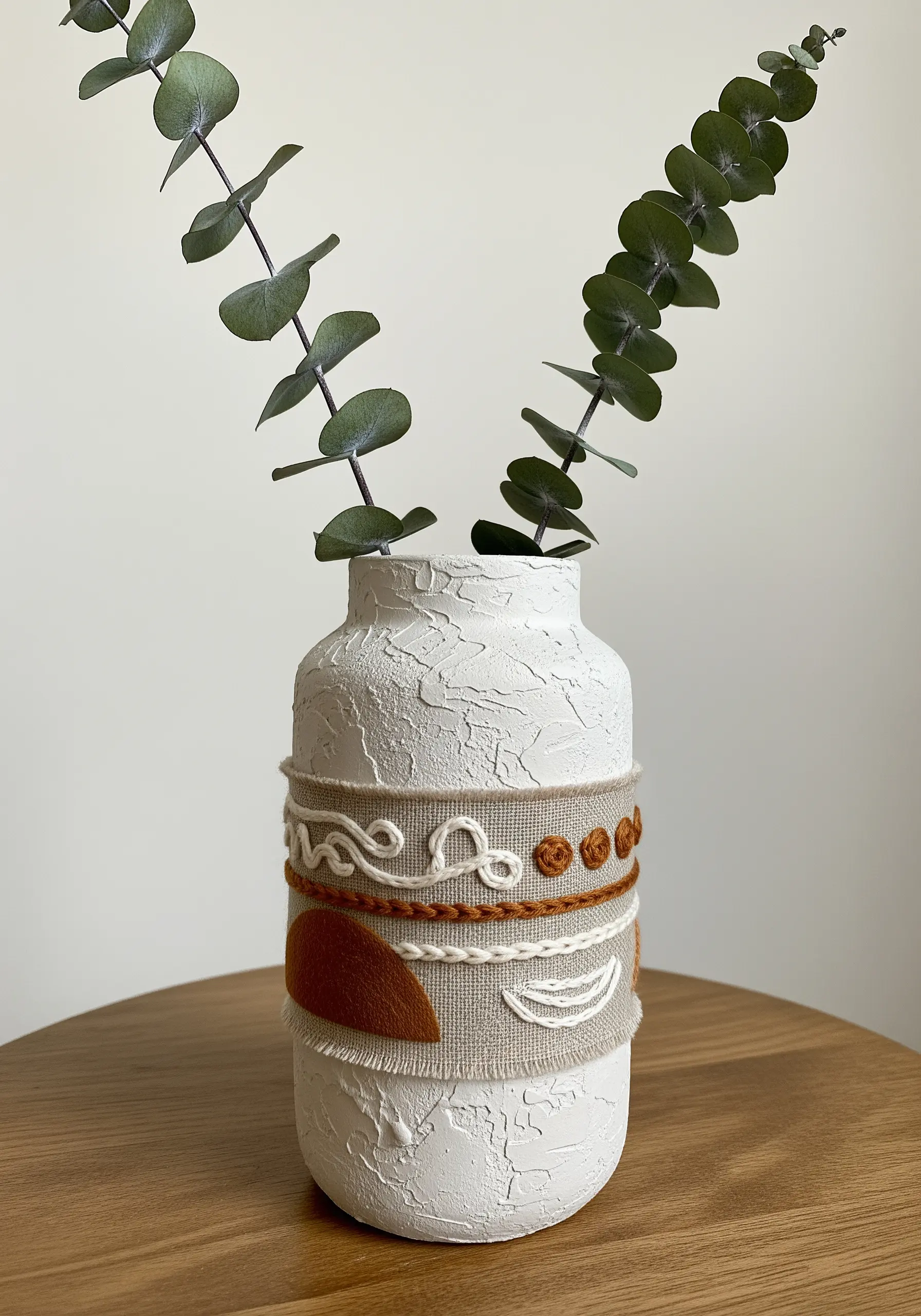 A white textured vase with a linen wrap embellished with yarn and embroidery in earthy tones.