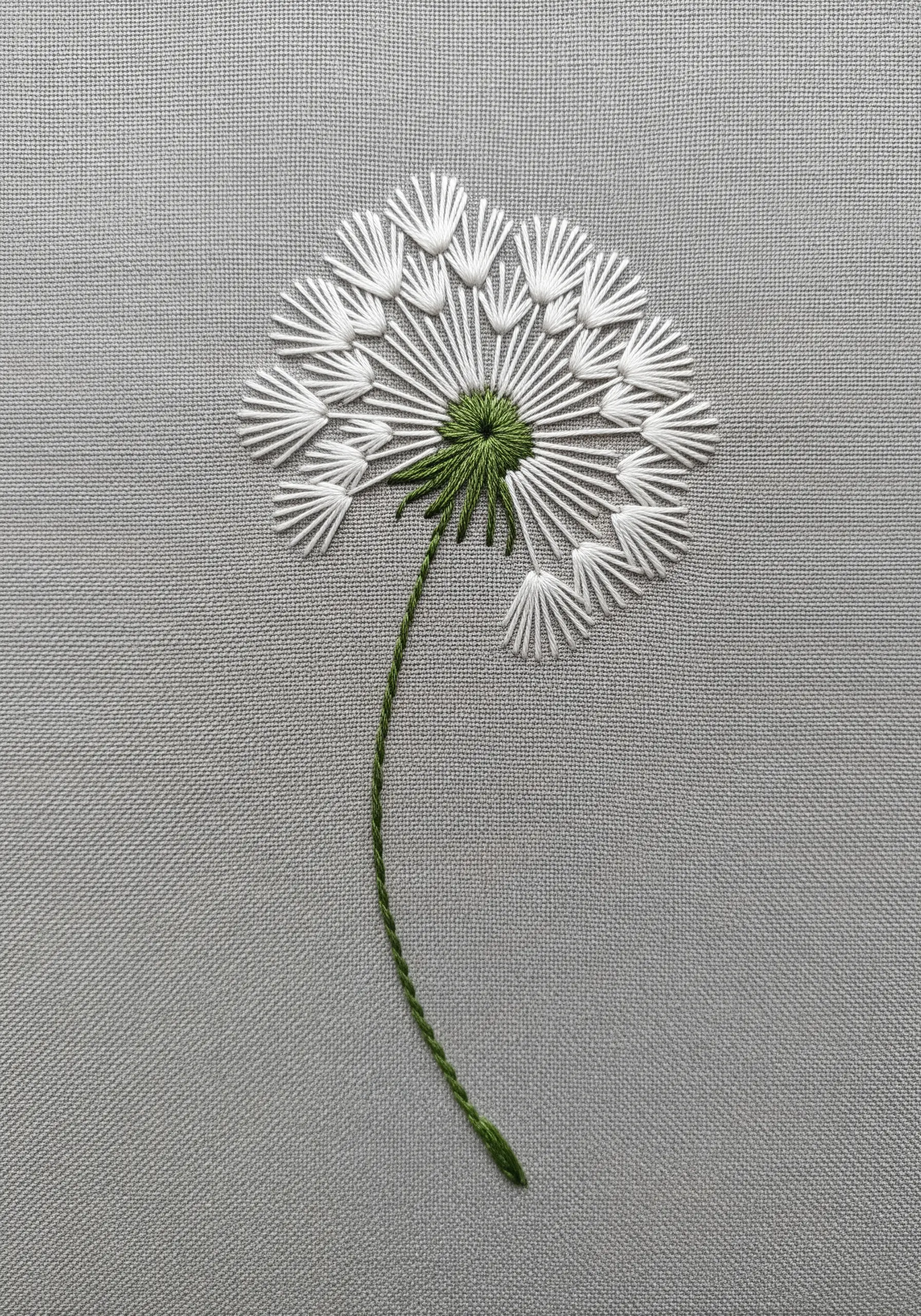 Dandelion seed head embroidered in white thread with a green stem on grey fabric.