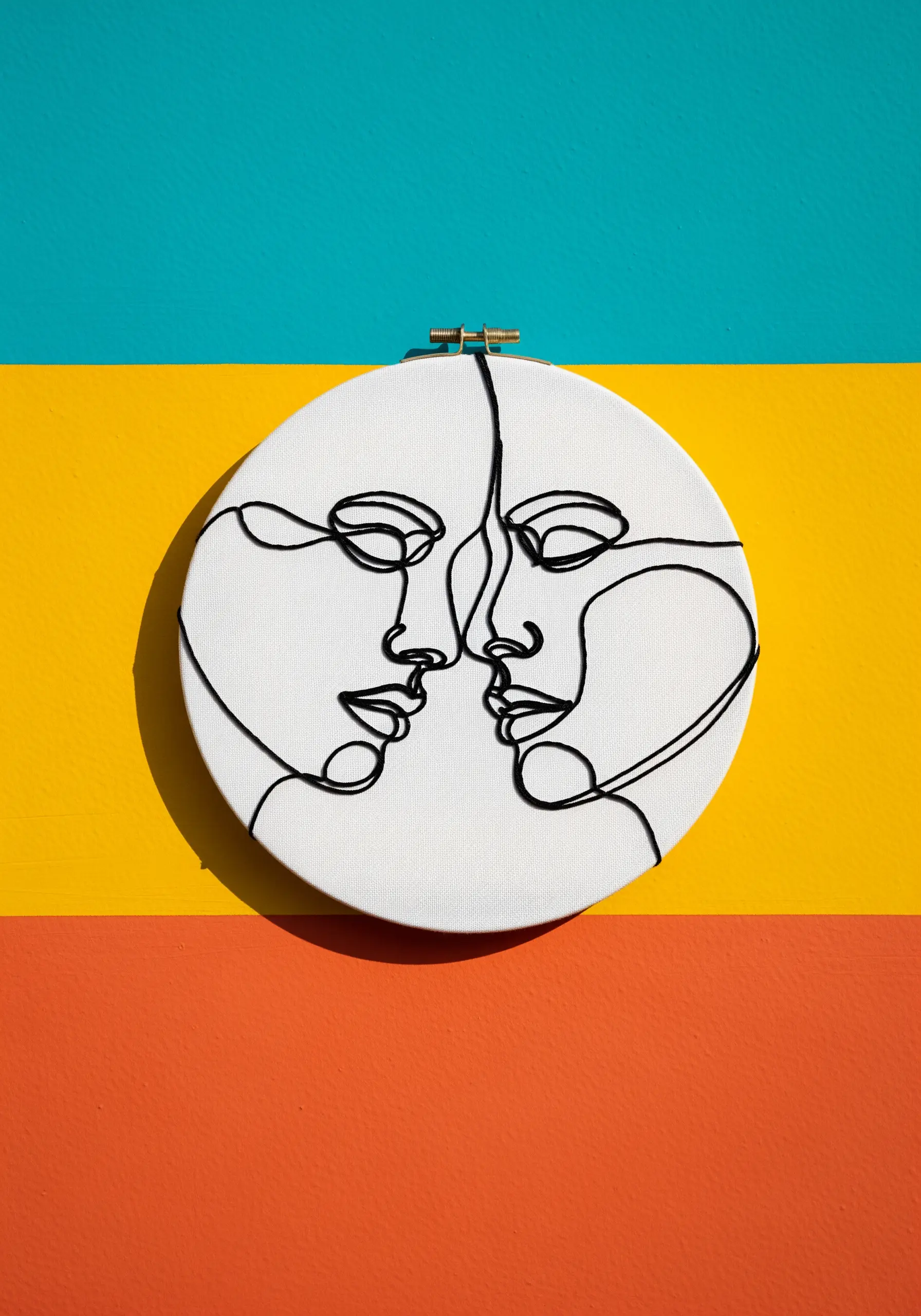 A one-line art portrait of two faces embroidered in black cord on a white hoop.