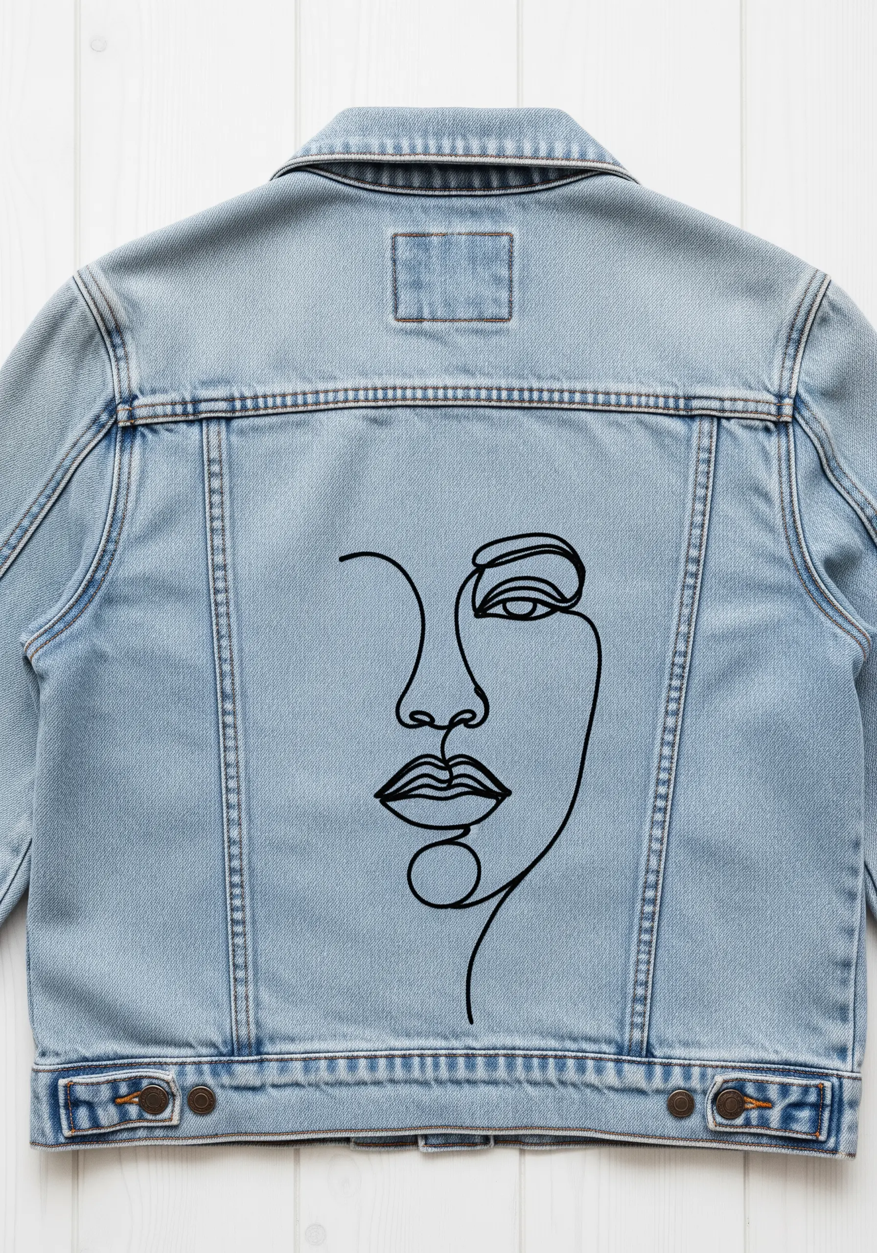 Minimalist black line-art portrait embroidered on the back of a light-wash denim jacket.