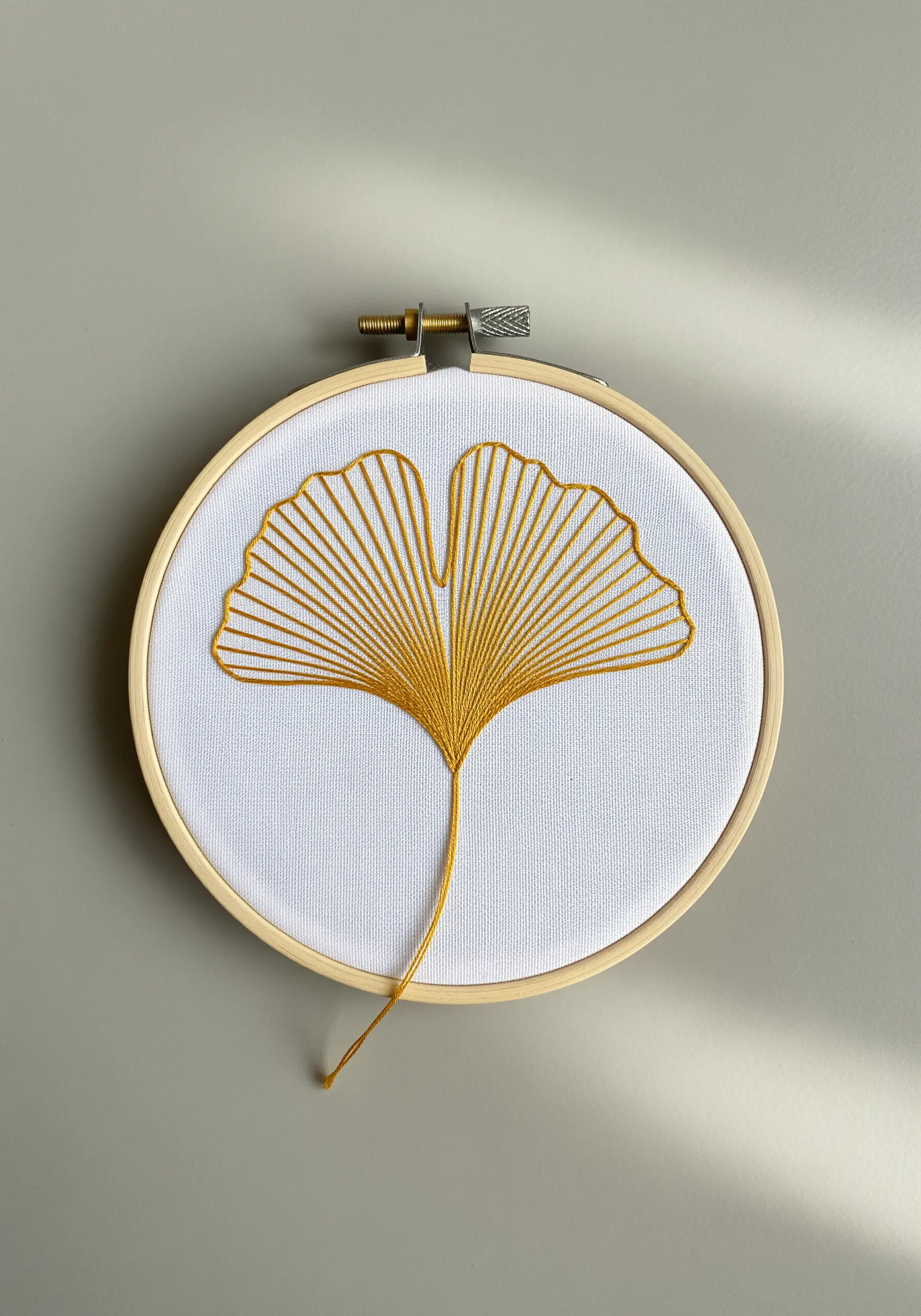 A golden-yellow ginkgo leaf embroidered with radiating straight stitches on white fabric.