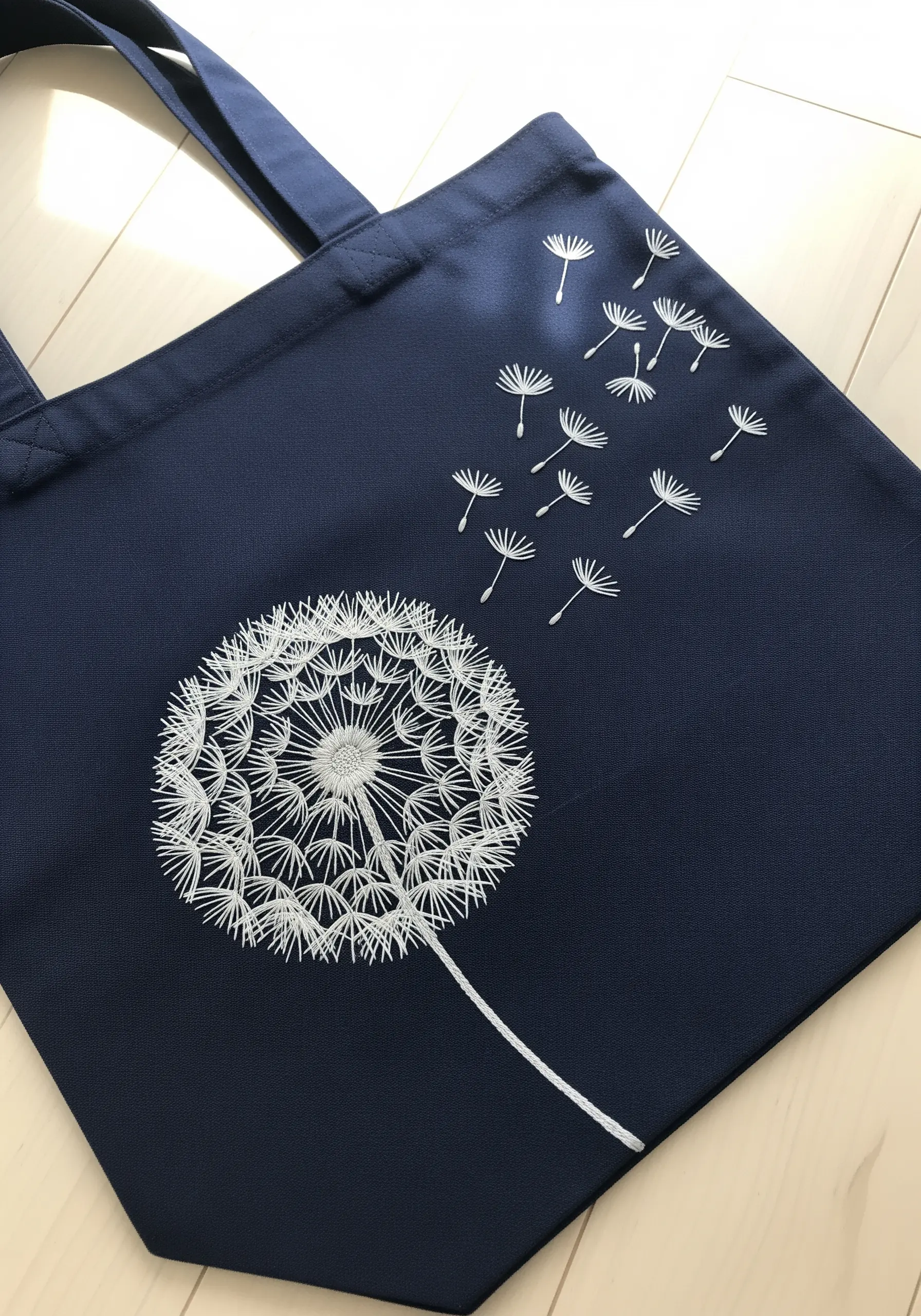 A single white embroidered dandelion with seeds blowing in the wind on a navy tote.