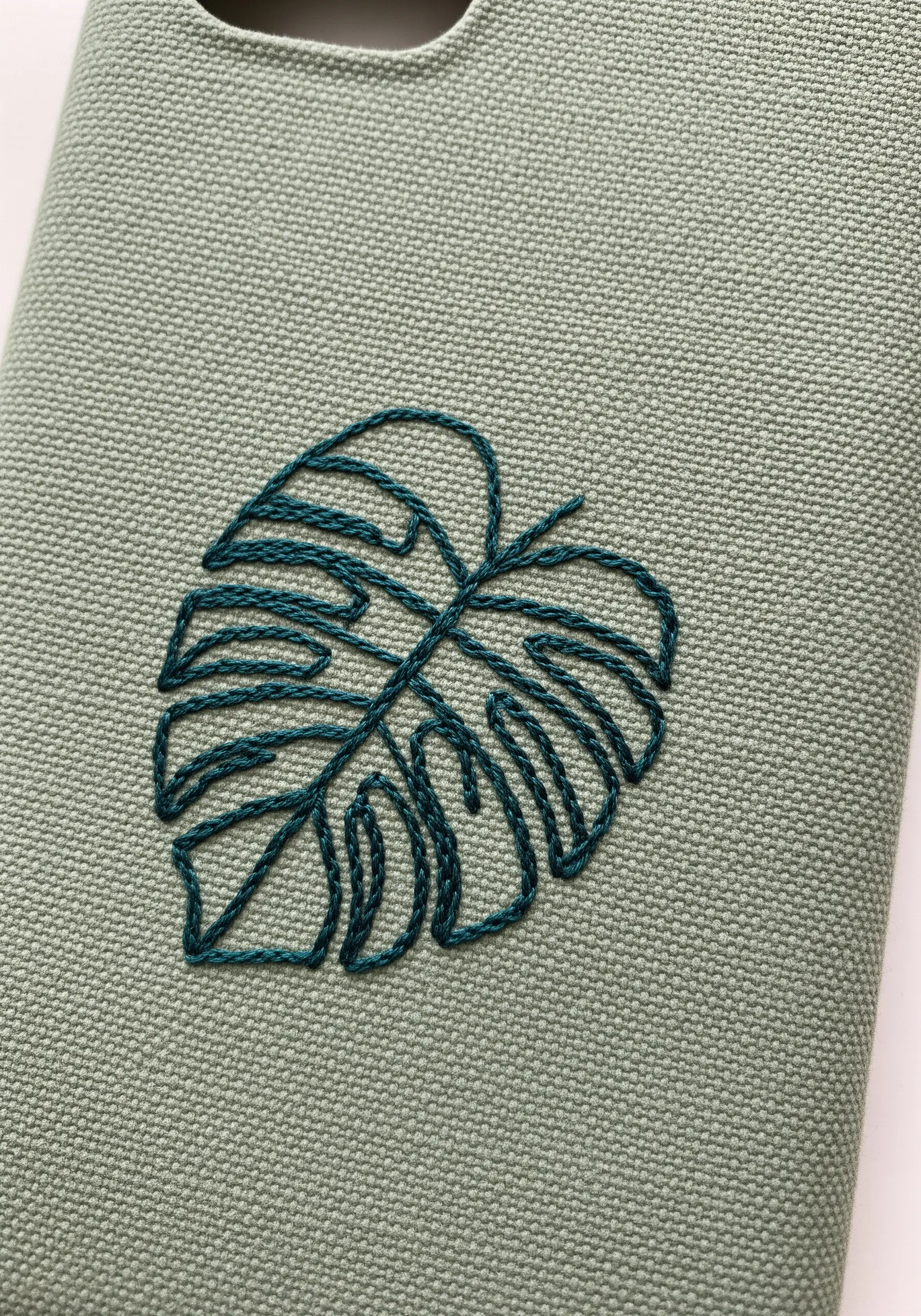 A single-color monstera leaf outline embroidered in dark teal on a sage green phone case.