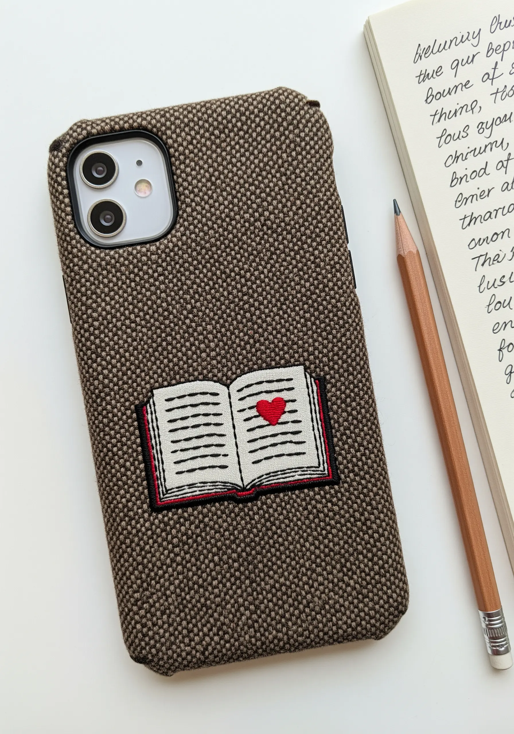 A small embroidered open book with a red heart on its page, stitched on a brown tweed case.